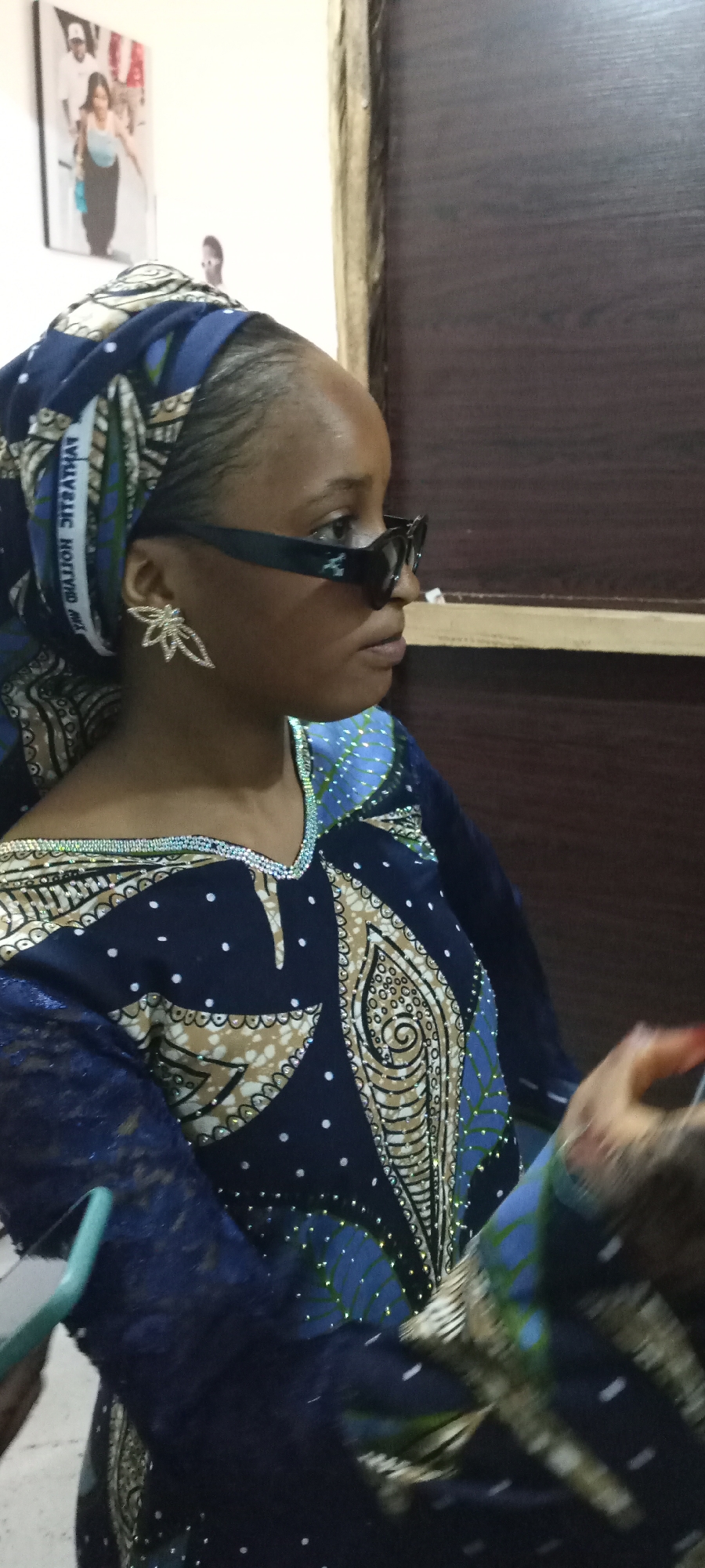 A medium shot depicts a dark-skinned person with a headscarf and sunglasses, viewed in profile facing right. The person has dark brown hair styled with braids peeking out from under a blue patterned headscarf. She wears dark-framed sunglasses with a subtle, reflective silver logo on the arm, and a shiny silver earring shaped like a stylized flower. Her attire consists of a dark blue top or dress decorated with lighter-colored patterns and small, scattered white dots. A trim of shiny beads lines the neckline.
The background includes a portion of a brown wooden door frame and a white wall with a framed picture of three people hanging on it. In the foreground, partially obscured, a person's hand and a light blue object are visible. The location is Unknown, Unknown.