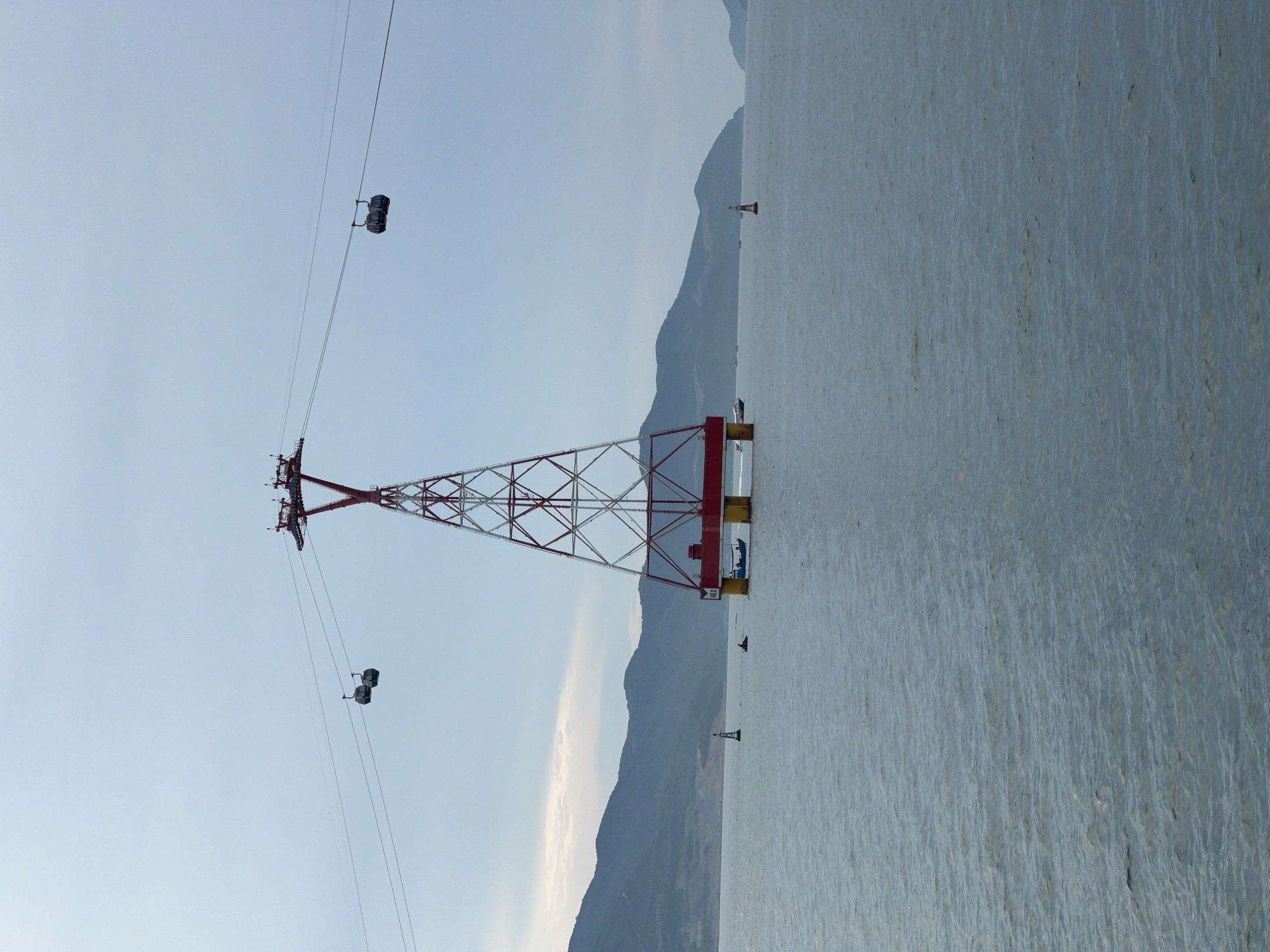 This is a vertical, eye-level, full shot of the Hon Tre Island cable car tower in Nha Trang, Vietnam. The tower is centrally located in the frame, supported by yellow pilings in the water. It is a red and white lattice structure with a red crossbar at the top, from which cable car lines extend to the left and right. Two cable cars are visible on the lines, one on the left and one on the right. The water is calm and gray, with small waves. In the distance, a chain of dark green mountains stretches across the horizon under a pale blue, overcast sky. Several small boats and buoys are scattered in the water. The scene is peaceful and expansive, conveying a sense of scale and isolation.