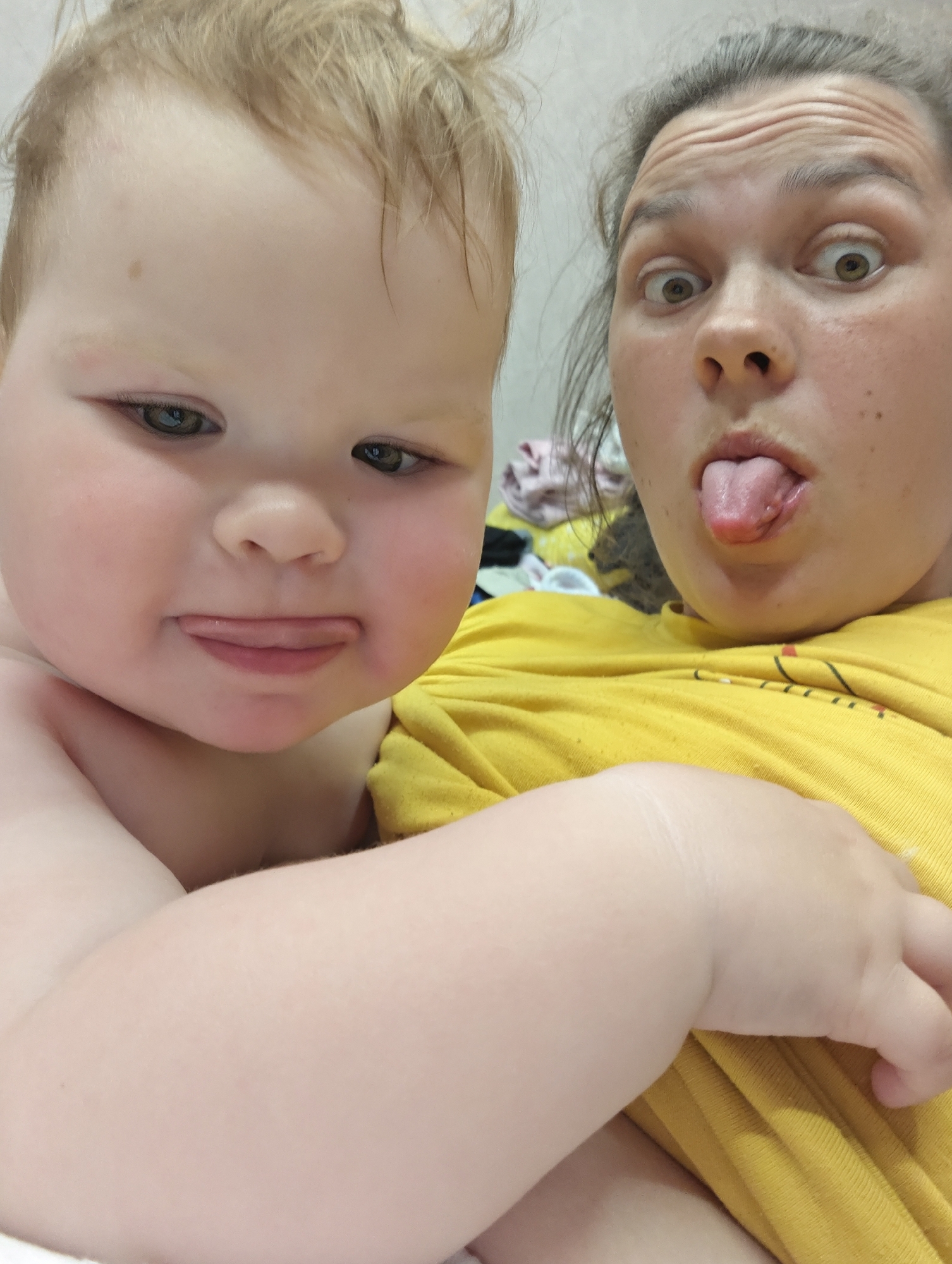 A close-up shot captures a woman and a toddler pulling funny faces. The toddler, positioned in the foreground on the left, is fair-skinned with light brown hair and is sticking out their tongue slightly. Their expression is relatively subdued. To the right, behind the toddler, a woman with her hair pulled back is making a surprised expression with wide eyes and her tongue extended out. She is wearing a yellow shirt. The background is a neutral wall. The location is not identifiable as the city of Chaikovsky, Russia from the image alone.