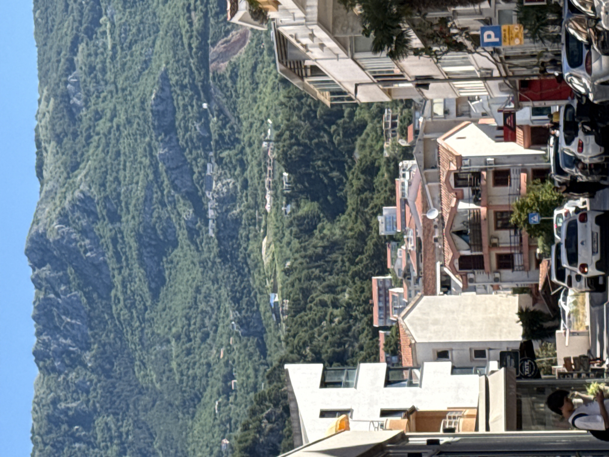 A vertical eye-level photograph shows a view from a street in the city of Budva, Montenegro. On the left is a view of a tree-covered mountain slope under a clear blue sky. A few buildings can be seen built into the side of the mountain in the distance.
On the right side of the photo is a street of buildings, mostly multi-story apartments and residences, with red-tiled roofs. A person walks past in the foreground of the image. Parked cars are seen lining the right side of the street, and a parking sign is affixed to a lightpost.