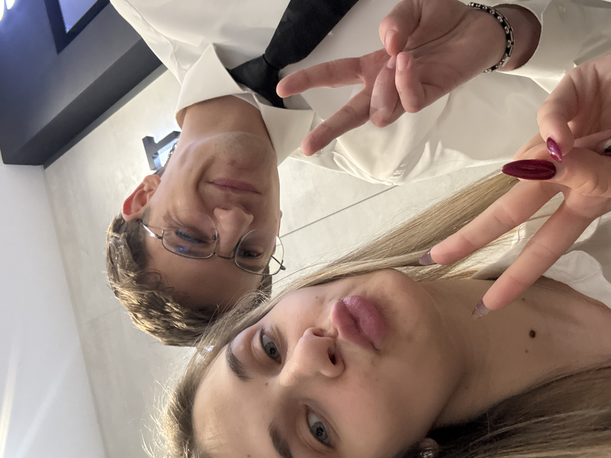 The image is a selfie taken indoors. Two individuals are visible; a male with short brown hair, glasses, and wearing a white shirt with a black tie, and a female with long blonde hair and manicured nails. Both are making a "peace" sign with their fingers. The male is wearing a bracelet. The background is blurred and offers no discernible details to determine the location in Sofia, Bulgaria.