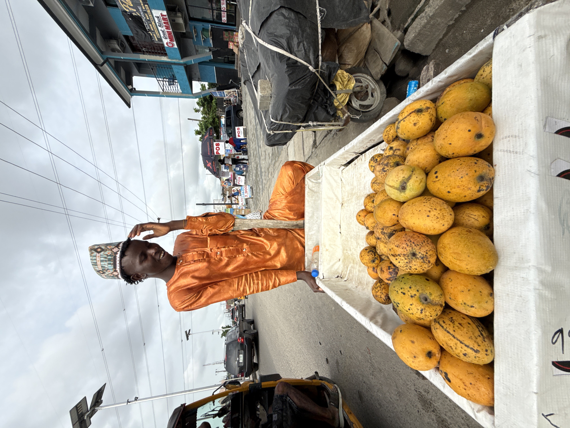 A street scene in Ado, Nigeria, features a man wearing a traditional patterned cap and an orange button-down shirt, standing by a cart laden with yellow mangoes. The cart's container is white. Behind the cart, another vehicle, covered with black material, is visible. Further back, the street shows parked cars and people by various signs, possibly indicating businesses or advertisements. The sky is overcast, and the foreground includes parts of a yellow tricycle taxi (Keke). A two-story blue and white building stands adjacent to the street. Overhead power lines are also visible.
