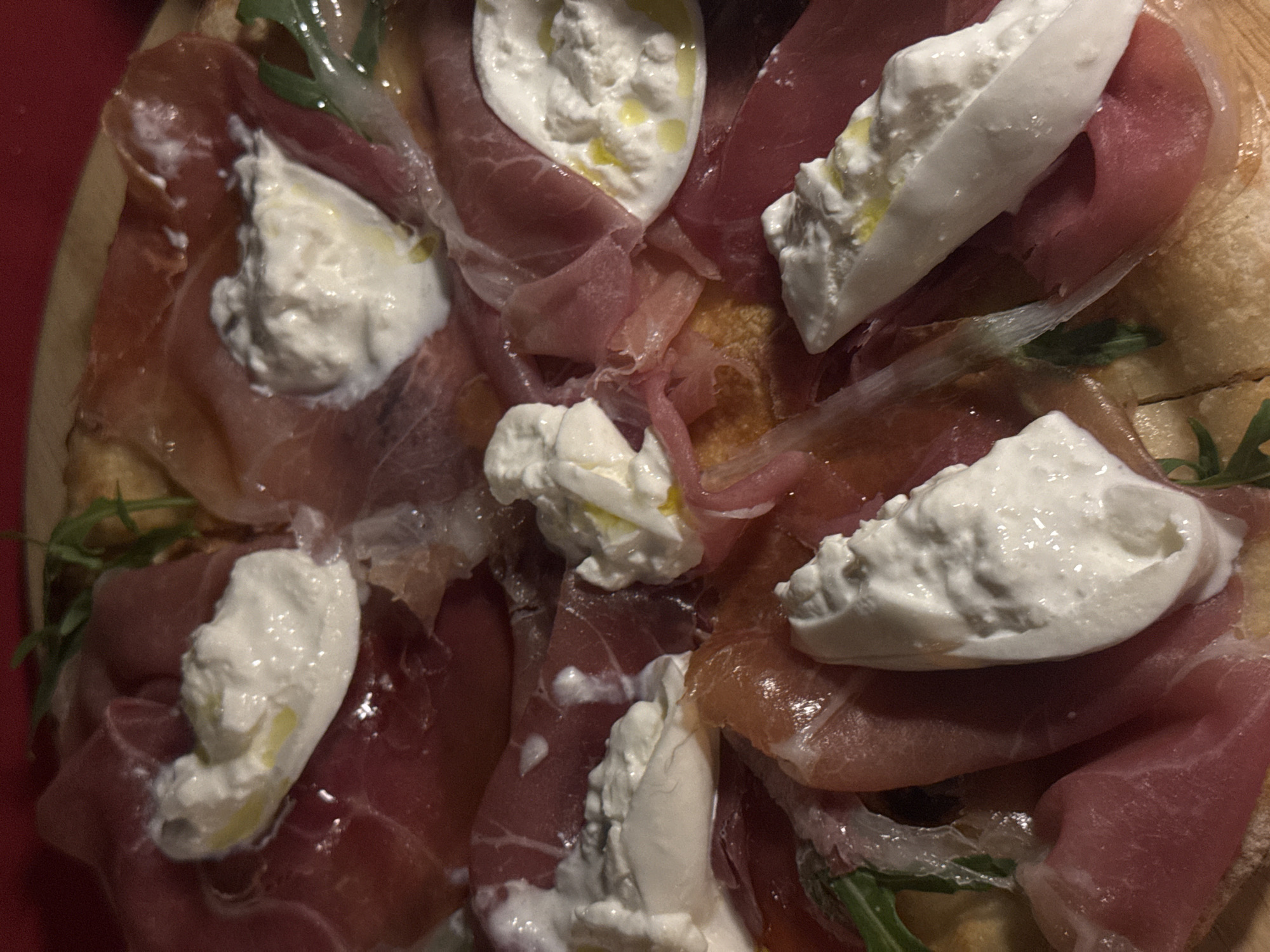 This is a close-up, overhead shot of a prosciutto and burrata pizza. The pizza crust is visible at the bottom, with thin slices of prosciutto draped over it. Dollops of fresh burrata cheese, drizzled with olive oil, are scattered across the top. A few sprigs of arugula are also visible. The lighting is dim and warm, creating a rich, appetizing atmosphere. It is impossible to determine the specific location within Chiang Mai City Municipality, Thailand based on this image alone.