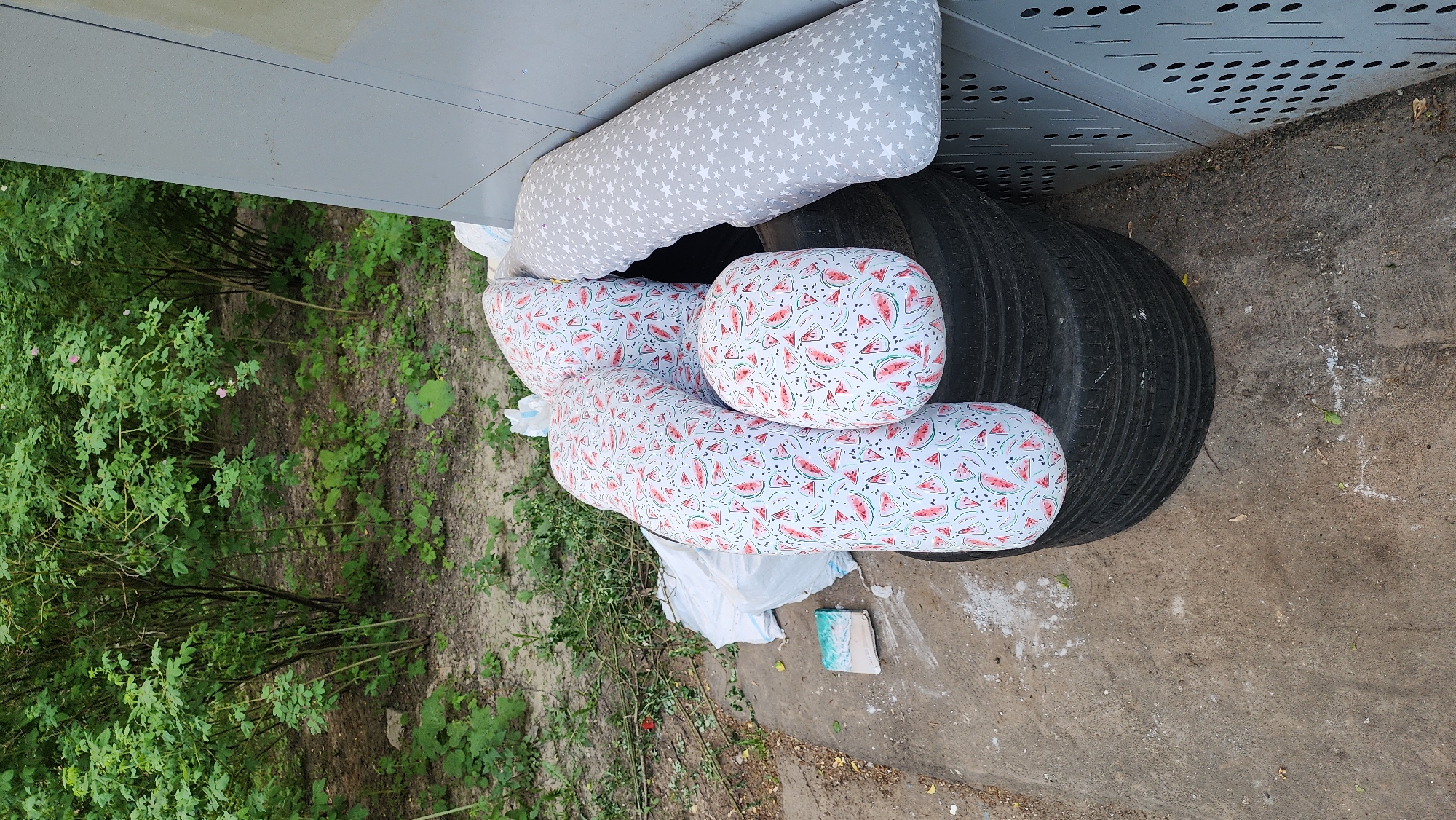 The image depicts a scene in an outdoor urban environment, likely a street or alleyway. A discarded stack of tires is visible, topped with several pillows. One pillow is patterned with grey stars, while others have a design featuring watermelons. The tires and pillows are positioned against a grey, perforated metal structure, possibly part of a building or utility installation. To the left of the objects, there is dense green vegetation, including bushes and possibly grass. The ground is a mix of concrete and dirt. There are discarded items, including what appears to be a small, rectangular object and some white trash bags or paper. There is no evidence to specifically suggest that this is Moscow, Russia. The scene appears to be a location where unwanted items have been left. No people are visible in the image.