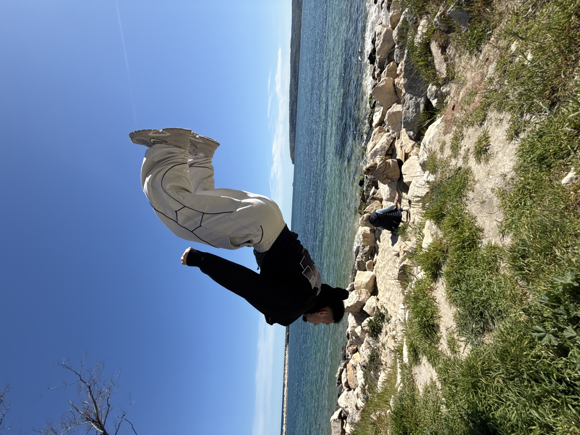 The image captures an athletic individual performing a dynamic backflip or acrobatic maneuver against a vibrant coastal backdrop under a clear blue sky. The main subject is airborne, suspended mid-flip, with their body curled. They are dressed in light-colored, baggy pants featuring dark geometric patterns and a black long-sleeved hooded top. The soles of their chunky, light-colored sneakers are prominently visible as they propel through the air.
In the background, a wide expanse of water, consistent with the Black Sea near Varna, Bulgaria, extends horizontally, exhibiting tranquil ripples and varying shades of turquoise and deep blue. A distant landmass or shoreline is faintly visible along the horizon. The immediate foreground consists of a rocky shore interspersed with patches of green grass. To the lower right, another person is partially visible, appearing to sit or kneel on the rocks close to the water, dressed in a light-colored jacket and dark attire.
The scene is illuminated by bright sunlight, suggesting a clear and sunny day with excellent visibility. A bare tree branch is subtly positioned in the lower-left corner against the brilliant blue sky. The photograph conveys a sense of energy, athleticism, and freedom within a picturesque natural setting. There is no visible text in the image.