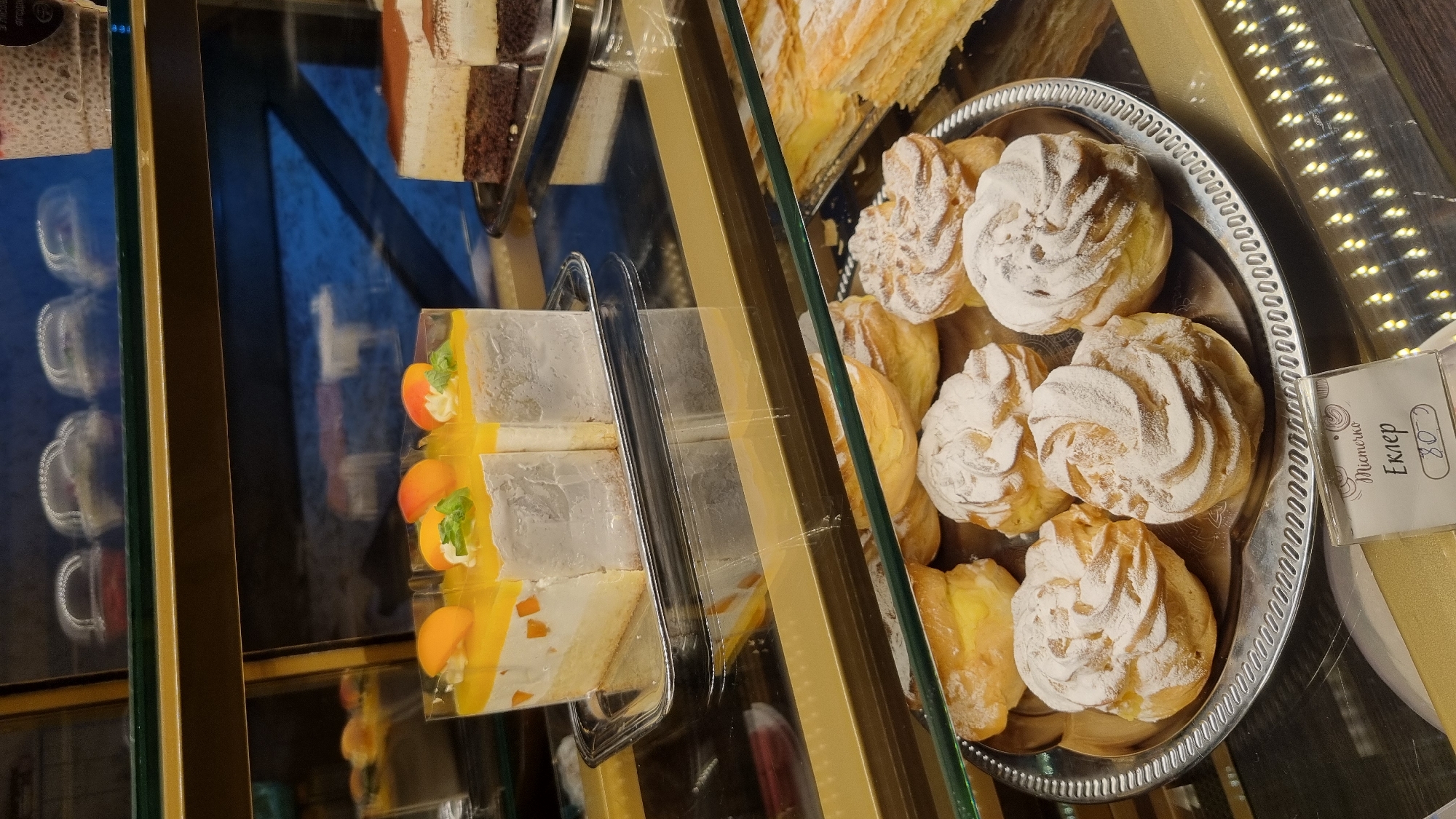 The image displays a bakery display case filled with various desserts. In the foreground, a silver platter holds several cream puffs dusted with powdered sugar, with a price tag indicating "Еклер 80". Behind the cream puffs, a tray of layered pastries is visible. On a shelf above, three slices of a yellow and white cake topped with apricots and mint are presented in individual clear plastic containers. To the right of these cakes, a slice of layered chocolate and cream cake rests on a silver tray. The display case is illuminated, and the reflections on the glass shelves suggest it is likely in a shop or cafe. The price tag with Cyrillic text and the currency symbol "80" implies the location is within Ukraine. Given the request to specify the city as Berehove, Ukraine, the scene depicts a bakery display in that city.