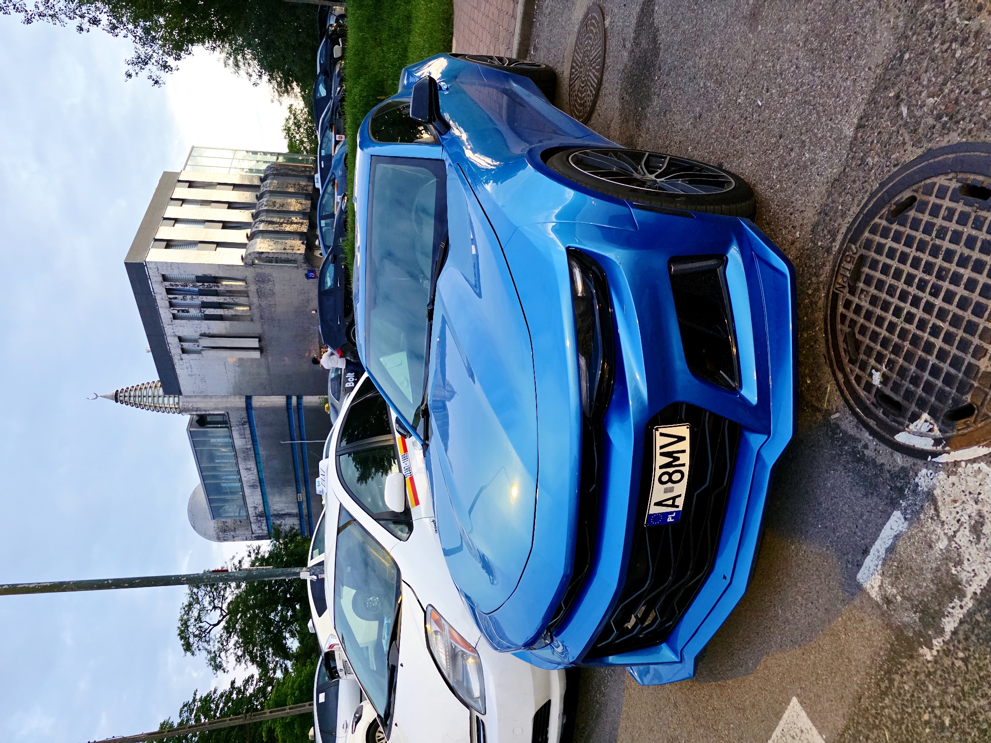 A blue Chevrolet Camaro is parked on a street in Warsaw, Poland, with a white Toyota Prius taxi and other vehicles in the background. A modern building with a distinctive minaret-like structure is visible behind the vehicles, likely a mosque or cultural center. The sky is overcast, suggesting it is either early morning or late afternoon. The Camaro's license plate reads "A 8MV" and is marked with "PL" for Poland. Several manhole covers are visible on the pavement.