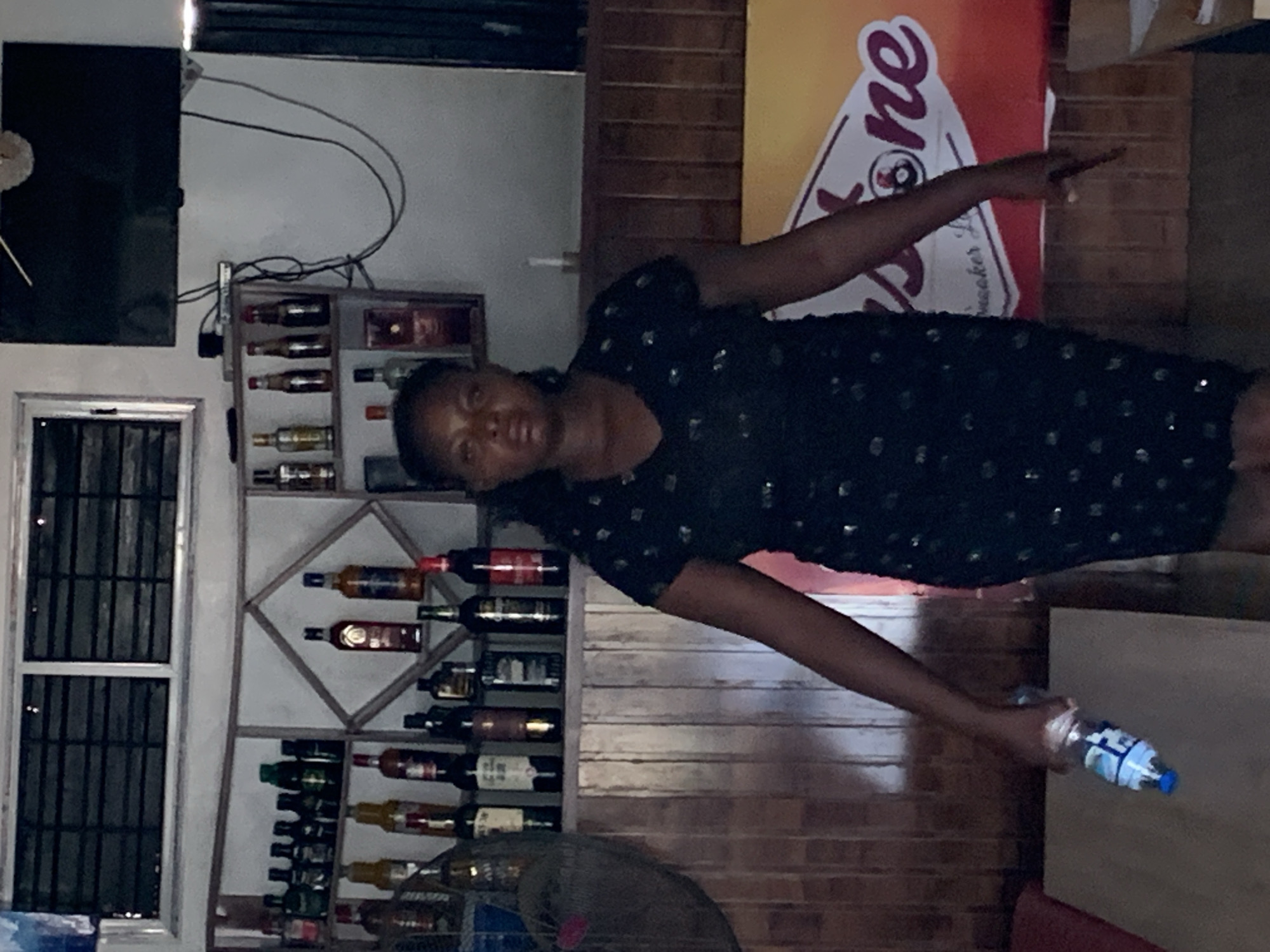A woman stands in what appears to be a bar or lounge in Jos, Nigeria. She is wearing a dark, sparkly dress and holding a bottle of water. Behind her, shelves are stocked with various bottles, likely alcoholic beverages. A television is mounted on the wall above the shelves. The environment appears to be indoors, with wooden paneling on the wall behind the woman and a large banner to her right.