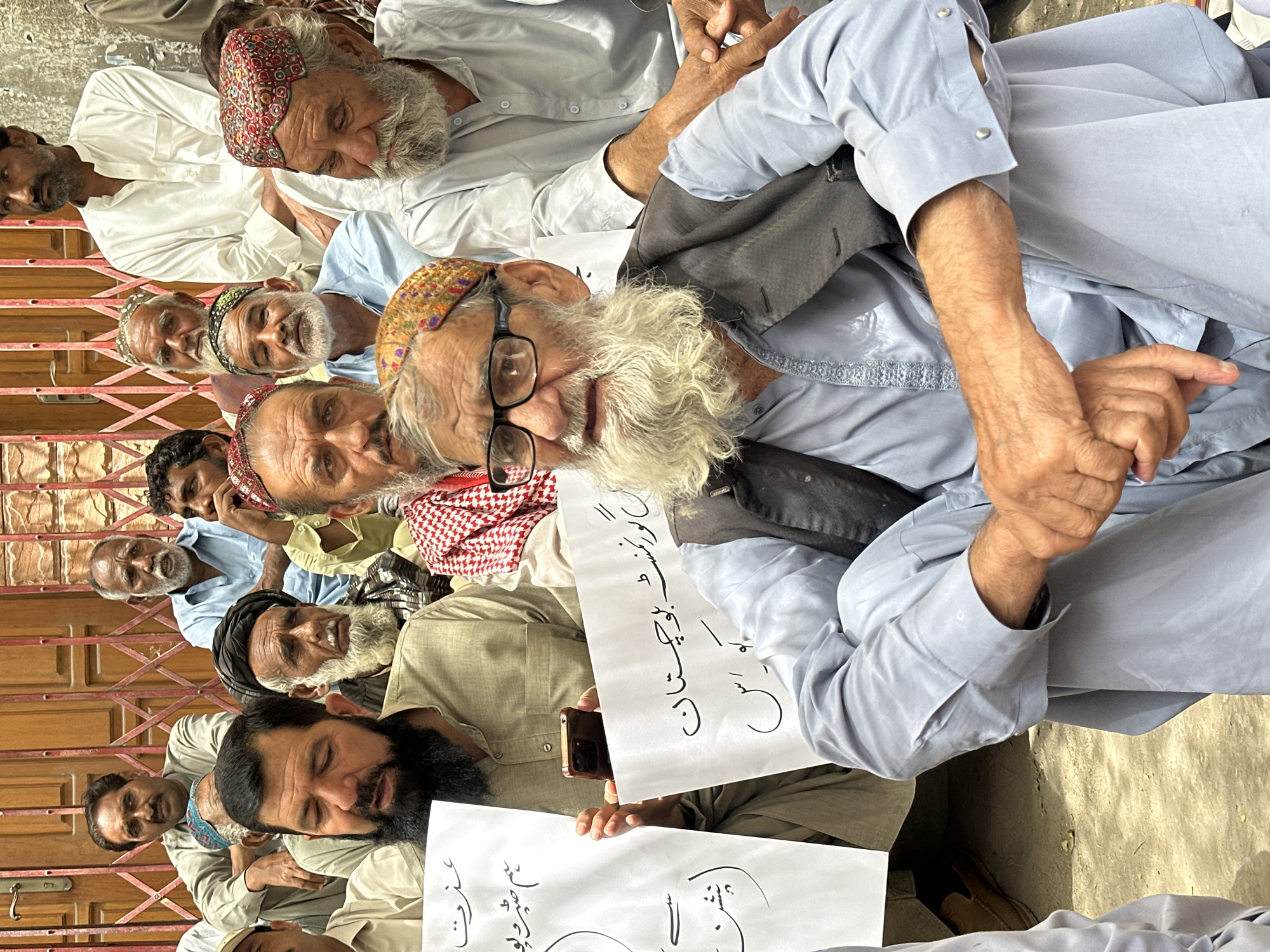 A group of men, most of whom appear to be of South Asian descent and are dressed in traditional clothing, are gathered outdoors in front of a building with a red metal gate. Several men are holding white sheets of paper with writing in Arabic script. One man in the foreground, wearing glasses and a grey vest over a light blue shirt, has a white beard and is looking towards the camera. The ground beneath them is dirt or gravel. The scene appears to be in Quetta City Tehsil, Pakistan, suggested by the presence of individuals and clothing typically seen in the region. The men seem to be engaged in some form of protest or gathering, indicated by the signs they are holding.