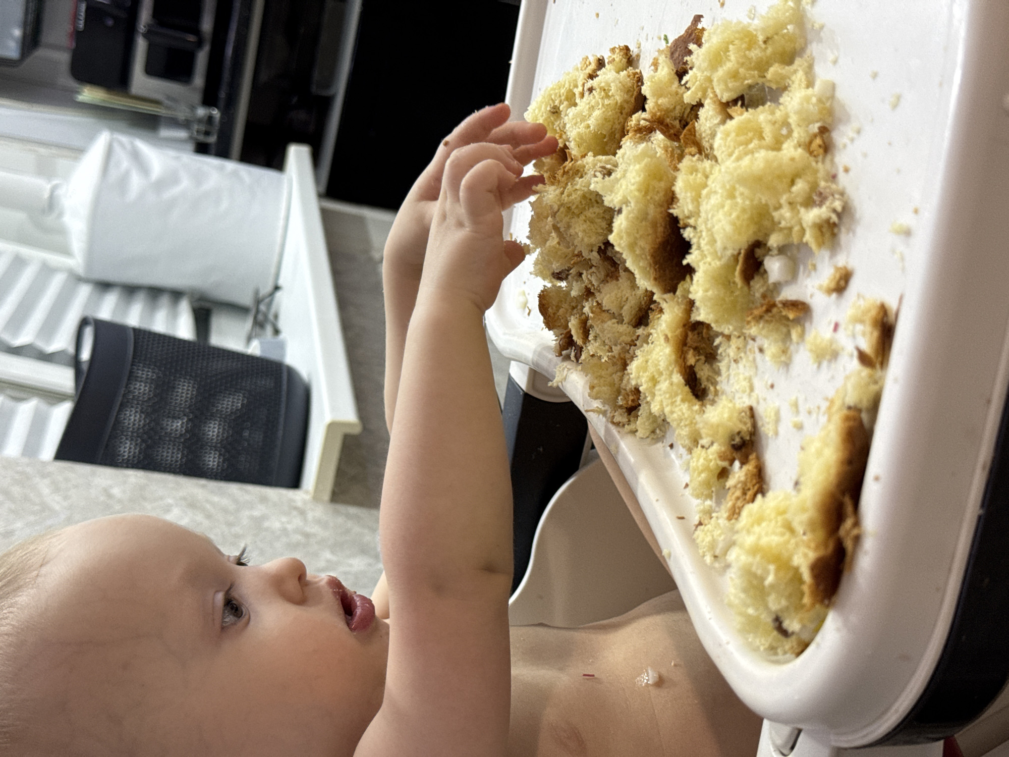 A baby is positioned in a highchair, reaching towards a tray covered with crumbled cake. The tray is white with a black edge. The baby has fair skin and is looking up at the cake with an open mouth. Parts of the baby's bare chest and arms are visible. The background includes a white object on a shelf, a black mesh object, and a white support structure. The location and city of Chaikovsky, Russia, are not discernible from this image.