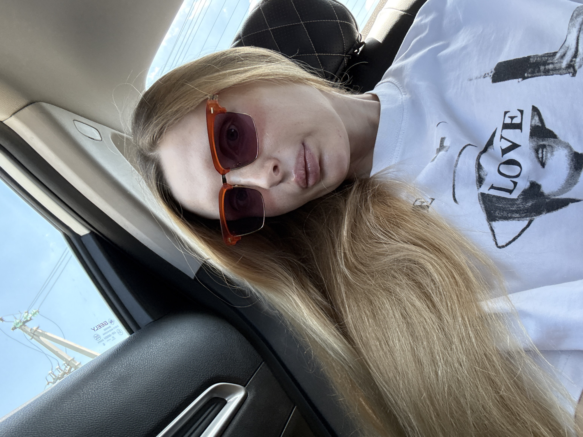 A young Caucasian woman with long blonde hair and orange-framed sunglasses is seated in the passenger seat of a vehicle, looking towards the camera. She is wearing a white t-shirt with a graphic print featuring the word "LOVE" overlaid on a person's face, along with an image of a tall building. The interior of the car is visible, including a black leather door panel and part of a headrest. The background through the car window shows a clear blue sky with utility poles and wires. The scene appears to be captured during daylight hours.