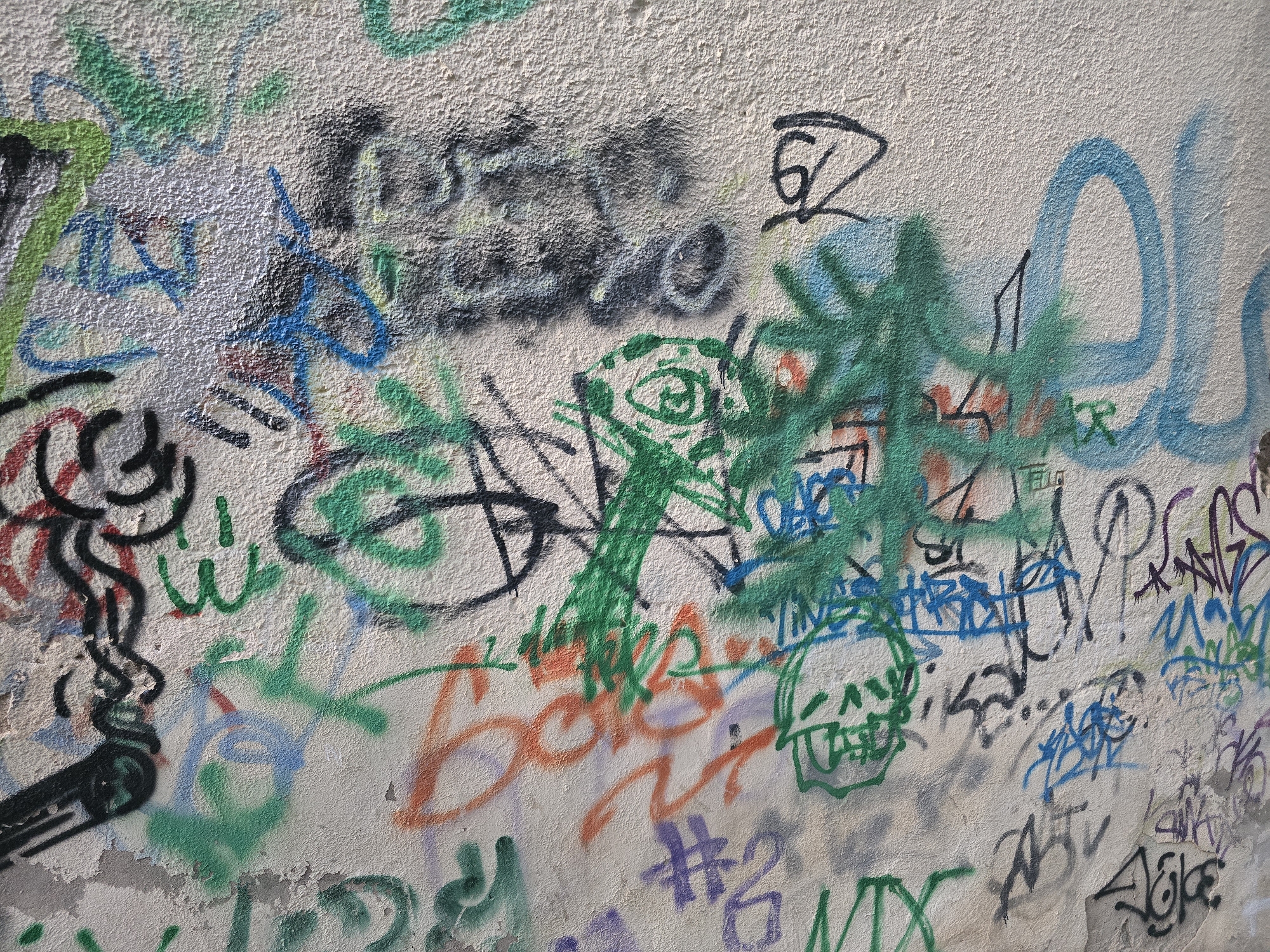 The image displays a close-up view of a graffiti-covered concrete wall. The graffiti is a mixture of spray-painted tags, scribbles, and figures in various colors including green, blue, black, red, orange, and purple. Some of the graffiti is abstract, while other parts appear to form letters, numbers, and stylized images, such as a smoking figure on the left and a skull-like figure in the center. The wall itself has a rough, textured surface and shows signs of wear and tear, with some peeling paint and chipped concrete. The lighting suggests it is daytime, casting subtle shadows on the wall. The specific location within the city of Jaén, Spain, cannot be determined from the visual information provided.