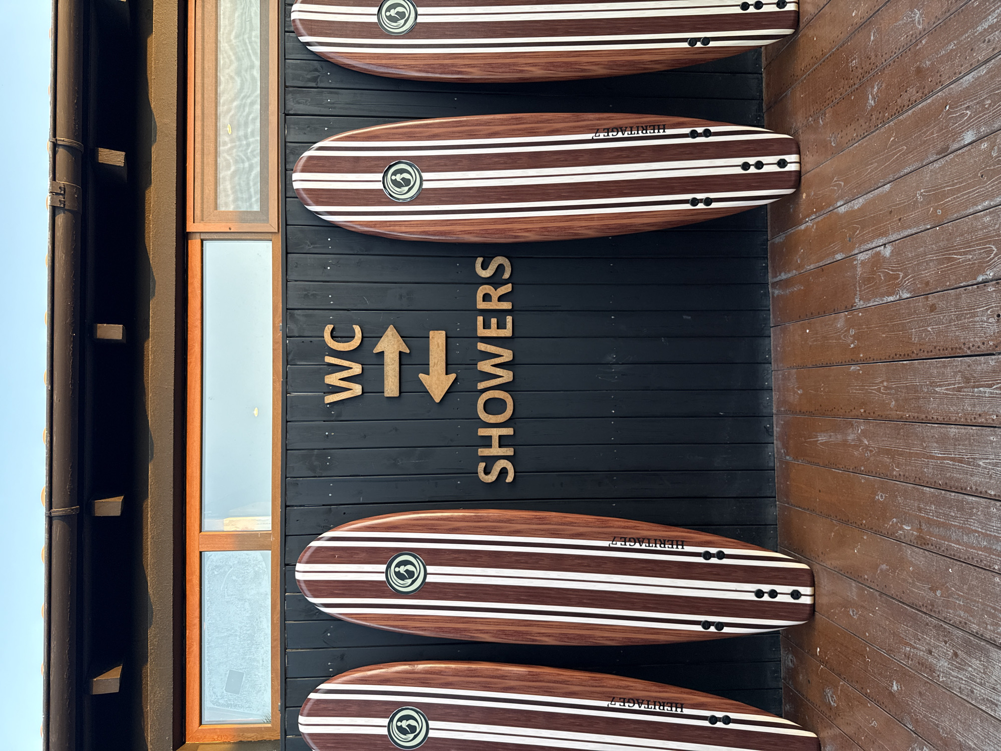 Four surfboards with a wooden finish and white stripes are mounted vertically on a black wooden wall. The surfboards are labeled "HERITAGE 7" and feature a circular logo with a wave design. Above the surfboards, wooden letters spell out "WC" and "SHOWERS", with an arrow pointing upwards between them, indicating directions to the facilities.
The black wooden wall is adjacent to a rustic brown wooden wall. To the left of the black wall are windows with wooden frames, suggesting an outdoor or semi-outdoor setting. The surfboards are arranged in a stacked pattern, with the top and bottom ones partially visible.
The image is taken from a slightly low angle, and the lighting suggests it could be daytime with natural light. The overall scene evokes a beach or surf-related establishment, possibly a surf shop or a facility near a beach. No people are visible in the image. The location context provided is Pallini Municipal Unit, Greece, which aligns with a coastal setting.