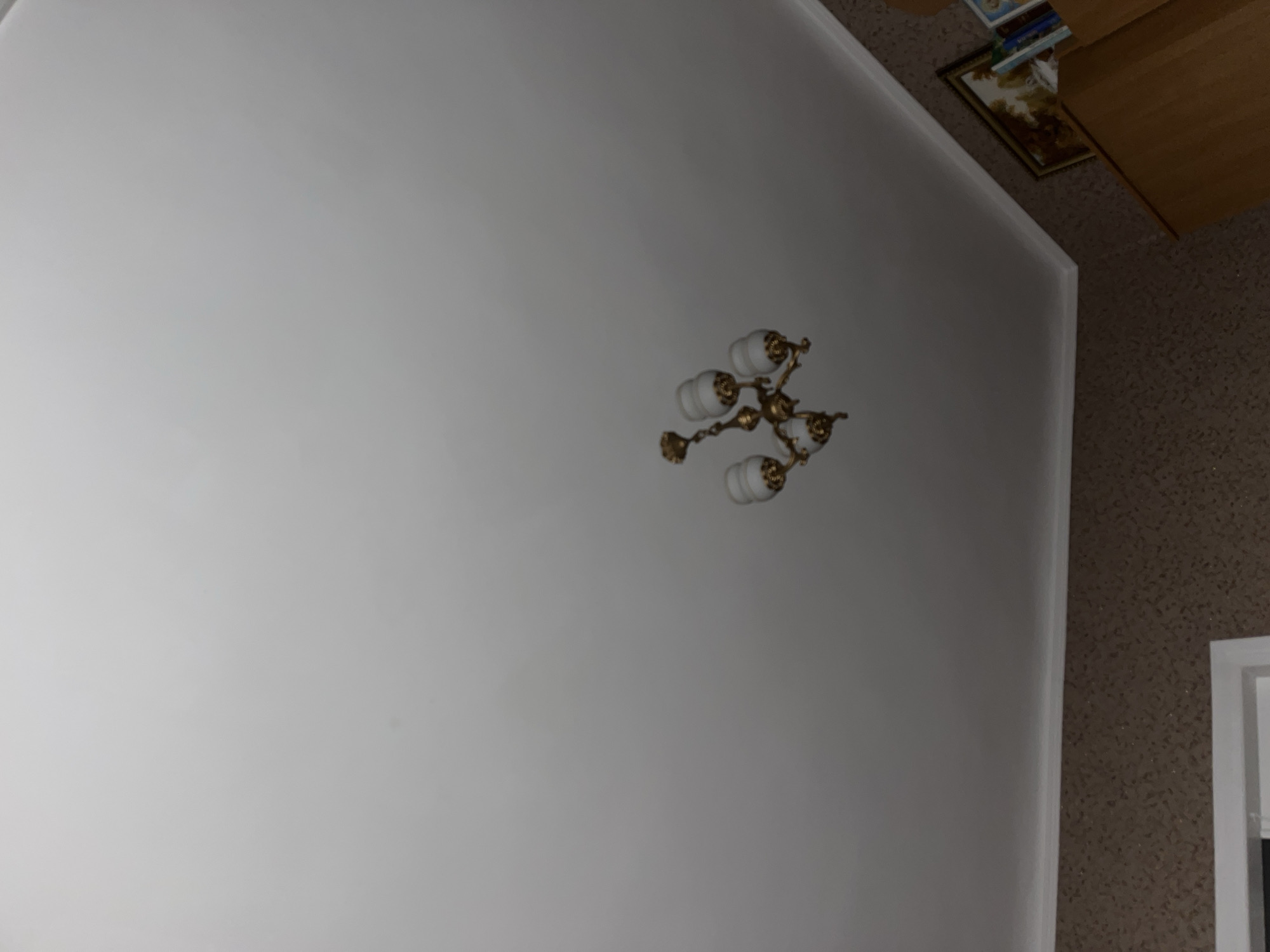 The image shows a ceiling with a vintage chandelier. The chandelier is made of gold-colored metal and has three white glass lamps. The ceiling is white and appears to be made of drywall. Below the ceiling, there is a wall with patterned wallpaper and a white baseboard. In the bottom right corner, a portion of a wooden cabinet and some framed pictures are visible. The scene is indoors, likely in a residential setting.