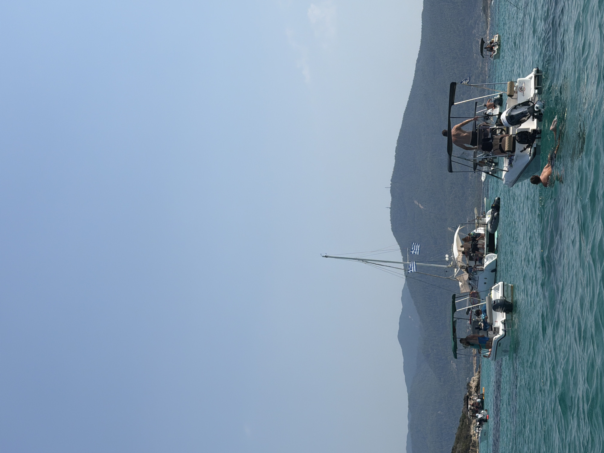 The image depicts a clear, sunny day at sea in an unspecified location in Greece. The turquoise water is calm with gentle ripples. In the foreground, a white speedboat with a black canopy is moored. Three people are visible on the boat: a man standing, wearing black shorts; a woman wearing a blue top and shorts; and a child in blue shorts. Another man is in the water swimming near the boat.
To the left, another white speedboat with a green canopy carries two people, a man and a woman, both casually dressed. Further in the background, a larger sailboat with two Greek flags flying from its mast is anchored. Several people are visible on the deck of the sailboat.
On the right side of the image, another white speedboat with a black canopy is visible, with a man standing at the helm. In the far background, a small white boat with a canopy is seen with a person on board. The shoreline is lined with green, forested hills that extend to a mountain range in the distant background. The sky is a clear, bright blue.