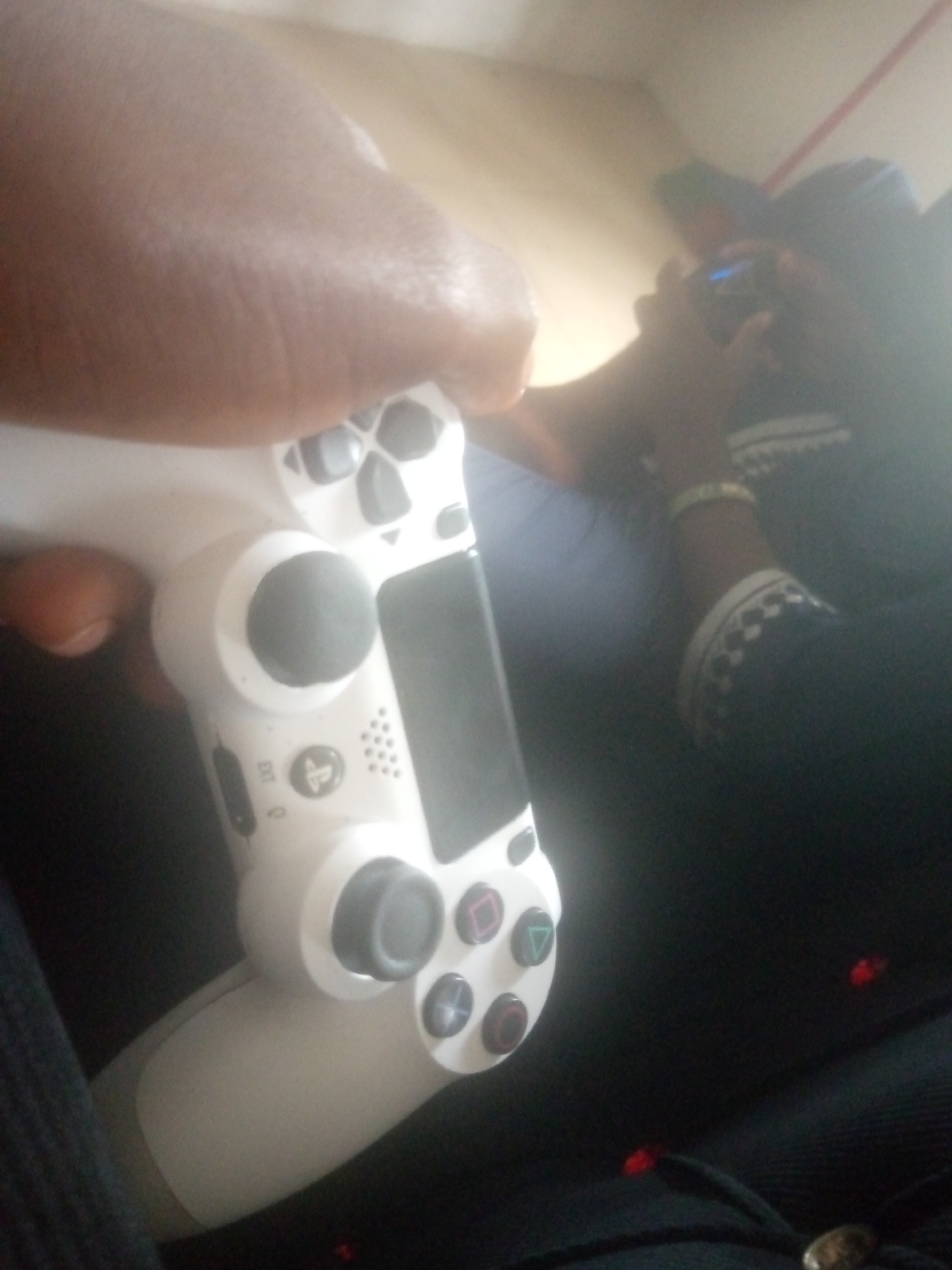 The image shows a person holding a white PlayStation 4 controller. The controller has black analog sticks and colorful face buttons, including a red circle, a blue cross, a green triangle, and a pink square. The person's hand is visible on the left side of the controller, and their thumb is positioned over the left analog stick. In the background, another person is holding a smartphone, and appears to be using it, with the screen glowing blue. The setting appears to be indoors, possibly a living room or bedroom, with soft lighting. The scene is casual and suggests a moment of leisure, with both individuals engaged in separate activities. The overall mood is relaxed. There is no visible text in the image.