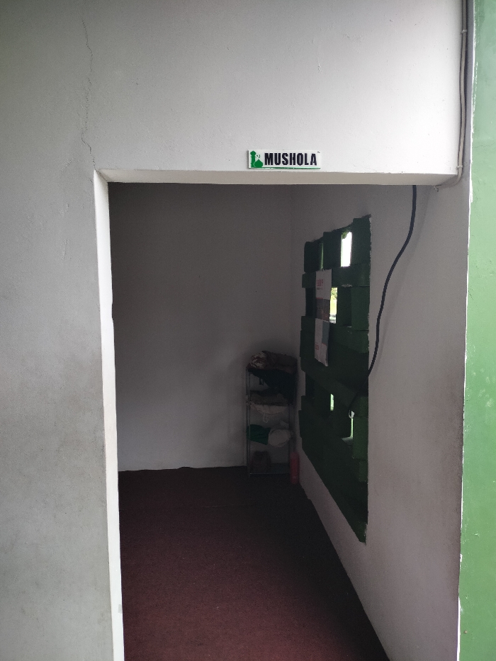The image displays the entrance to a small, empty room, identified as a "MUSHOLA" by a sign above the doorway. The sign is white with green text and a stylized mosque or minaret icon. The entrance itself is a simple rectangular opening in a white painted wall.
To the left of the entrance, an exterior wall, possibly concrete or stucco, is light gray and shows a distinct vertical crack. On the far right edge of the frame, a vertical section of a green structure or wall is visible, suggesting an exterior element.
Inside the room, the walls are painted white, and the floor is covered with a dark red or brown carpet. On the back wall, a simple, multi-tiered metal shelving unit holds various folded items, which appear to be fabrics or clothes, along with some round objects resembling plates or bowls. A small red object, possibly a fire extinguisher, stands on the floor near the shelf.
The right interior wall features a section painted dark green, composed of horizontal slats or panels with small rectangular openings, through which natural light appears to enter. A black electrical cable runs vertically down this green section. The overall lighting suggests it is daytime. The room is unoccupied and appears to be a functional, modest prayer space, consistent with the "MUSHOLA" designation in the Dramaga, Indonesia context.