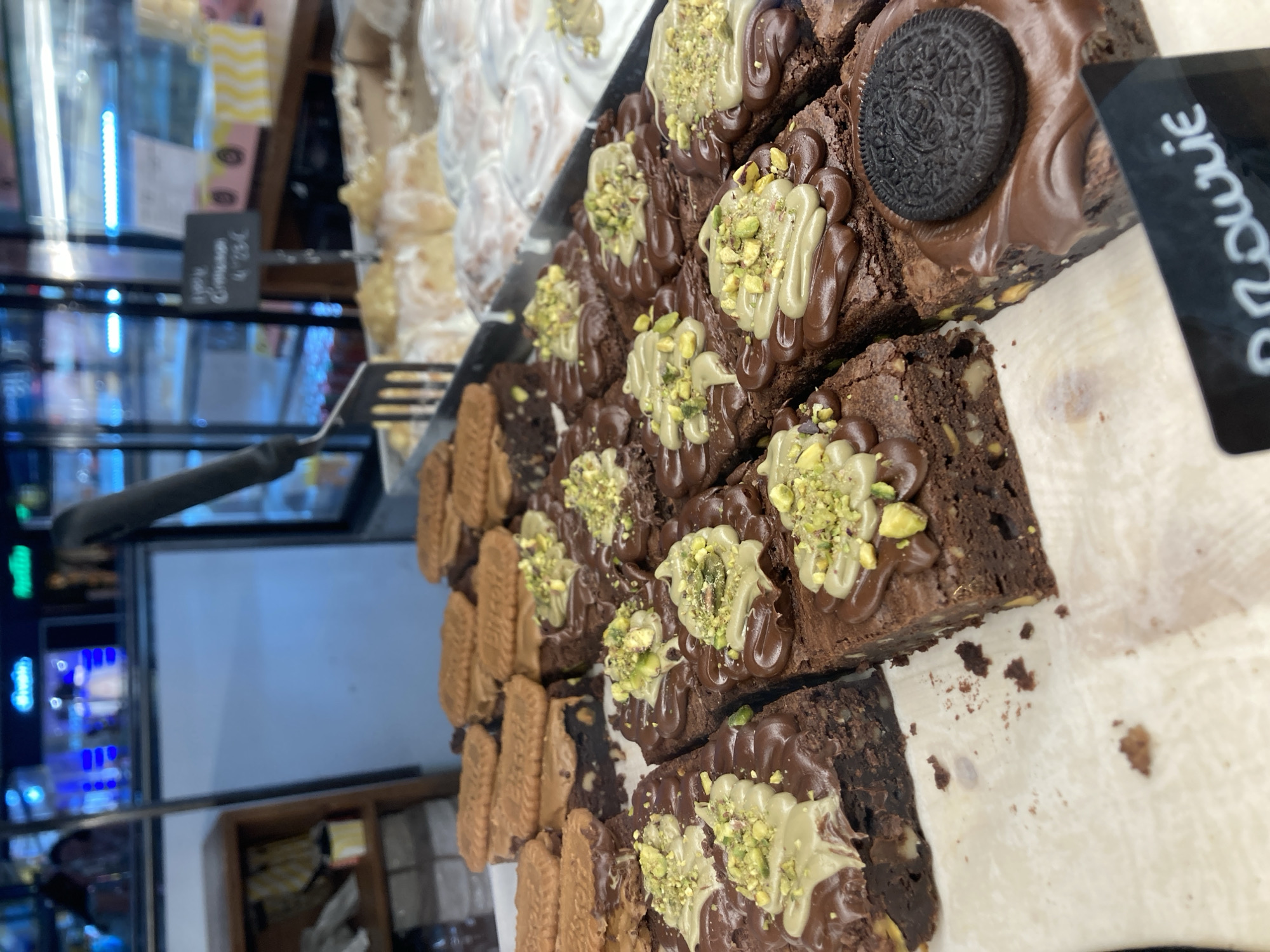 In Barcelona, Spain, a close-up, eye-level shot showcases a display of freshly baked brownies. The foreground is dominated by rows of rich, dark chocolate brownies, each adorned with a swirl of lighter colored frosting and a generous sprinkle of chopped pistachios. One brownie in the front right corner is topped with a dark chocolate frosting and a whole Oreo cookie. In the background, other baked goods are visible, including what appear to be cinnamon rolls with white icing. Behind the baked goods, a refrigerated display case with blue lighting and various products can be seen, along with a wooden shelf holding what appear to be packaged goods. A metal serving utensil rests on the edge of the display. A small black sign with white lettering, reading "Brownie", is visible in the lower right corner of the image.