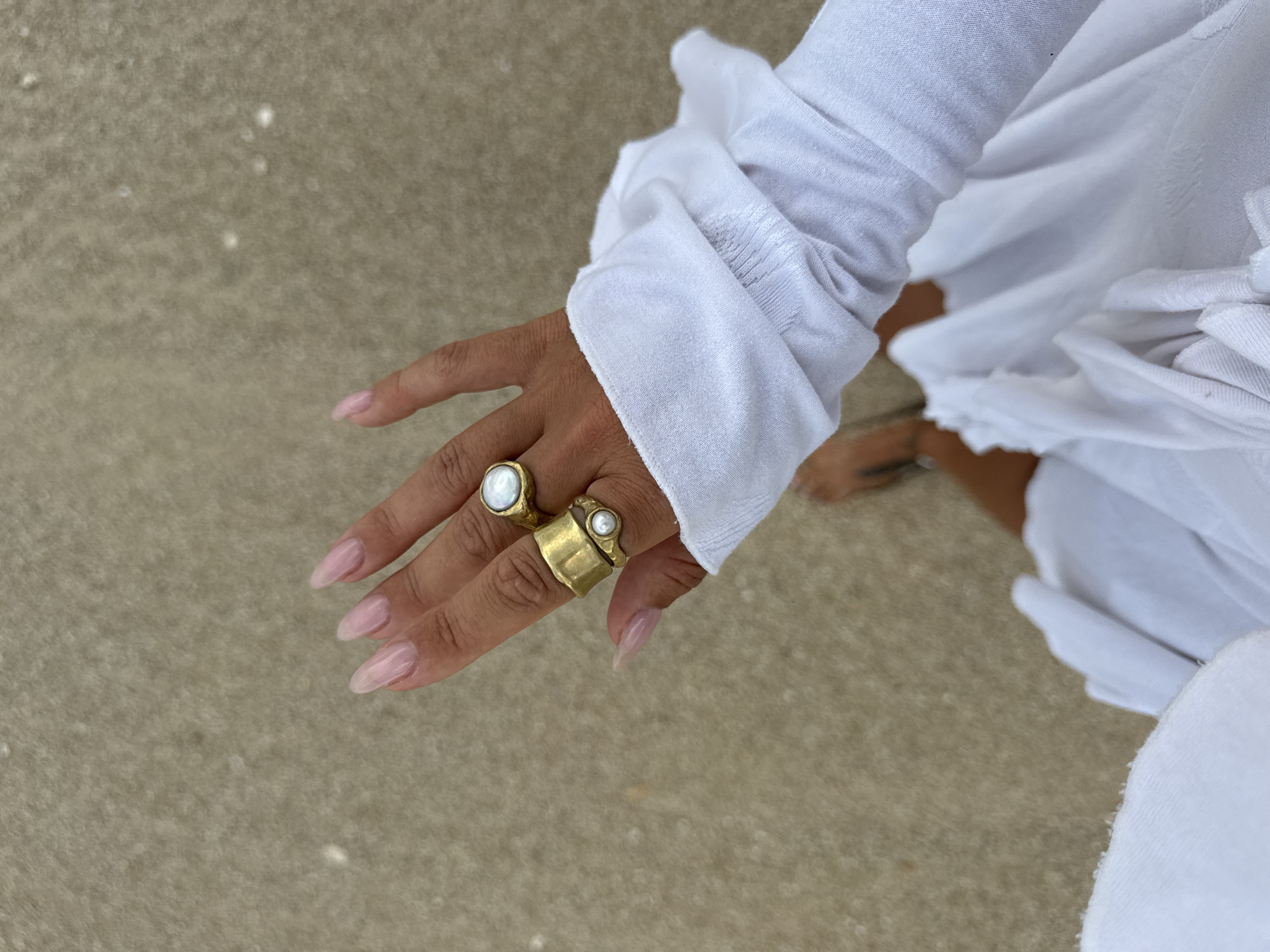 A person's left hand, adorned with two gold rings featuring pearls, is held up with the palm facing the viewer, against a backdrop of a sandy surface. The hand has long, light pink manicured nails. The person is wearing a white long-sleeved shirt, with the cuff pushed up, revealing a portion of their arm. The scene is set on a beach in Kuta Selatan, Indonesia.