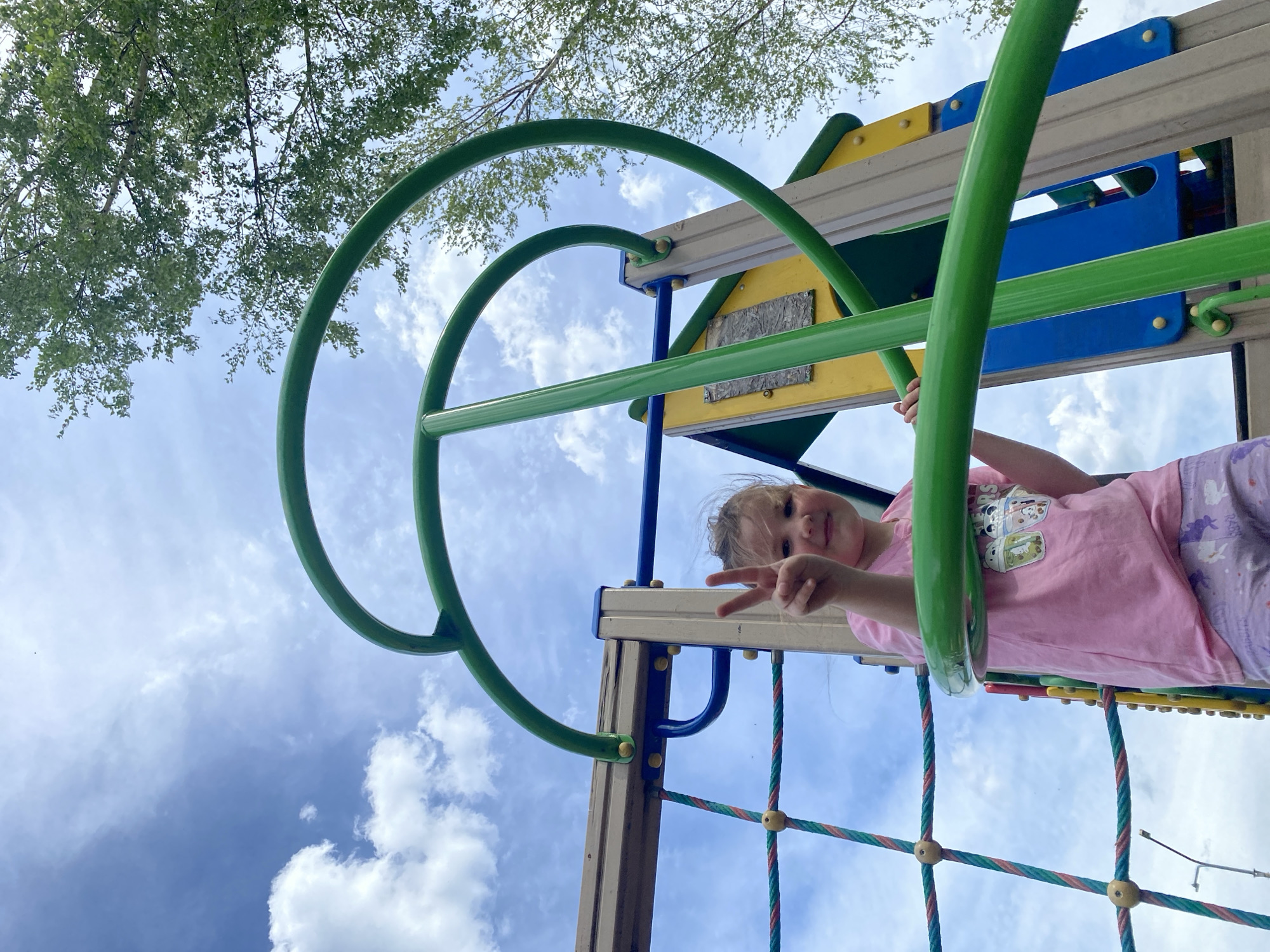 A young girl, angled upwards, is playing on a playground structure. She is wearing a pink t-shirt and lilac patterned leggings. She is holding onto a green, circular monkey bar and making a "peace" sign with her fingers. The playground structure is multi-colored, with elements of yellow, blue, and beige. A rope climbing net is visible. Trees and the sky, filled with white clouds, are in the background. The location is Chaikovsky, Russia.