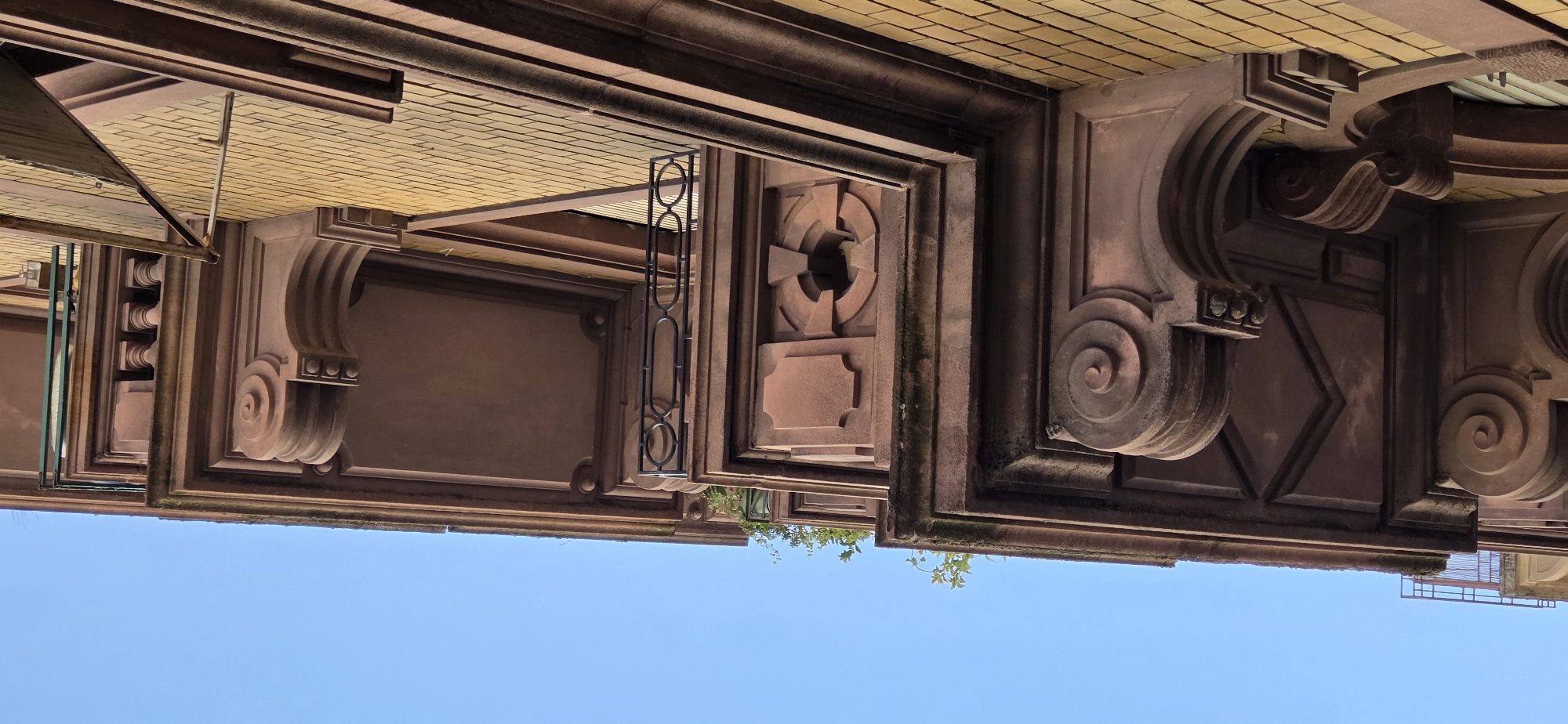 The image shows an ornate architectural detail of a building's facade against a clear blue sky. The perspective is from below, looking upwards, and the image is rotated 90 degrees counter-clockwise. The facade features elements of neoclassical or art deco design, characterized by carved stonework in a reddish-brown hue. Prominent decorative elements include large, voluted scrolls, geometric patterns, and rectangular panels. Above the stonework, a section of yellow brickwork is visible, suggesting the upper part of the building. A thin, dark metal railing or grate is also integrated into the design in one section. Some greenery, likely ivy or vines, is creeping up the facade in a few places, hinting at an outdoor setting. The bright sky suggests it is daytime, likely sunny. The architectural style and the presence of yellow brickwork are common in many European cities, and the provided location context points to Strasbourg, France. There is no visible text in the image. The scene focuses solely on the architectural details and the sky, with no people or specific activities depicted.