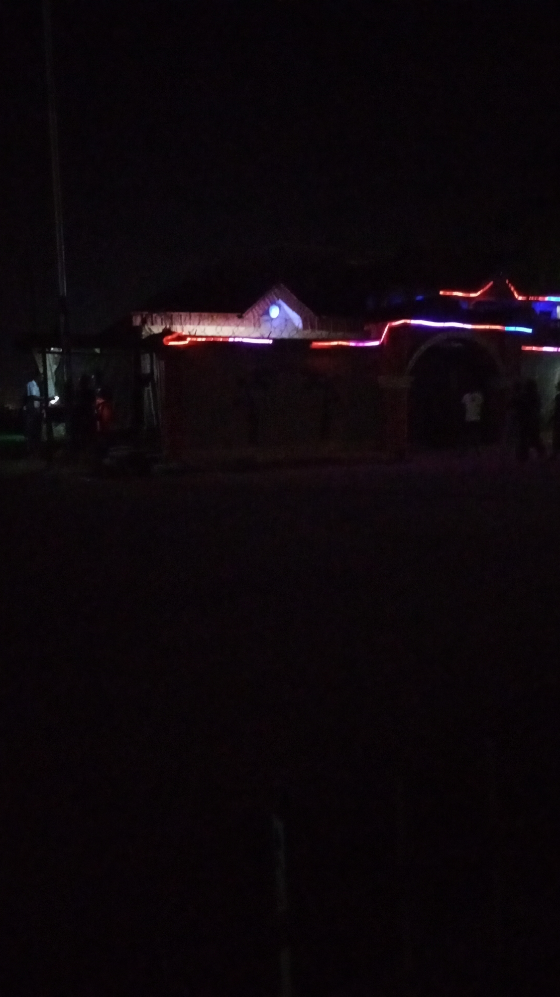 The image shows a building at night, likely in an urban environment given the lack of stars and bright ambient lighting. The building is decorated with colorful string lights in red, blue, and purple along the roofline and possibly around an archway. The building appears to be made of a dark material, possibly brick or stone, and has a prominent archway or entrance on the right side. There are indistinct figures in front of the building to the left and right sides of the main door. The foreground is a dark street or open space. A tall, possibly metal pole is visible on the far left. A house to the top right has a pointy roof that is colored white. The location appears to be Unknown, Unknown.