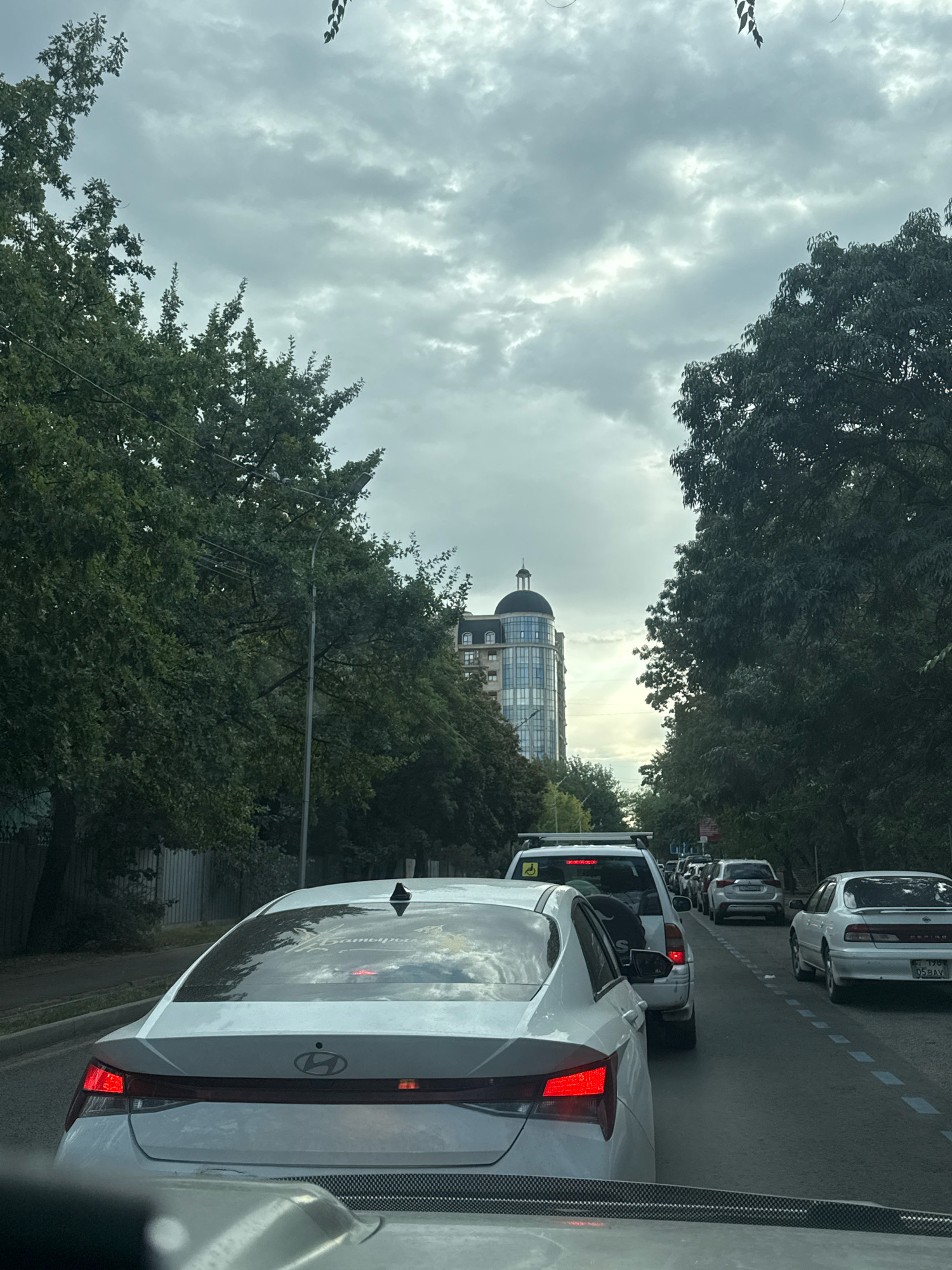 A view from a vehicle in traffic on a city street in Almaty, Kazakhstan, under a cloudy sky. The foreground shows the rear of a white sedan. In front of it are several other vehicles, including a white SUV with a yellow accessibility symbol on its rear door, a white compact SUV, and other cars further down the road. On the right side of the street, a white sedan with a license plate reading "05 BAV 176" is visible. Lush green trees line both sides of the street, with a modern, glass-fronted building topped with a dome visible in the distance through a gap in the trees. The road is paved, and dashed white lines mark the lanes.