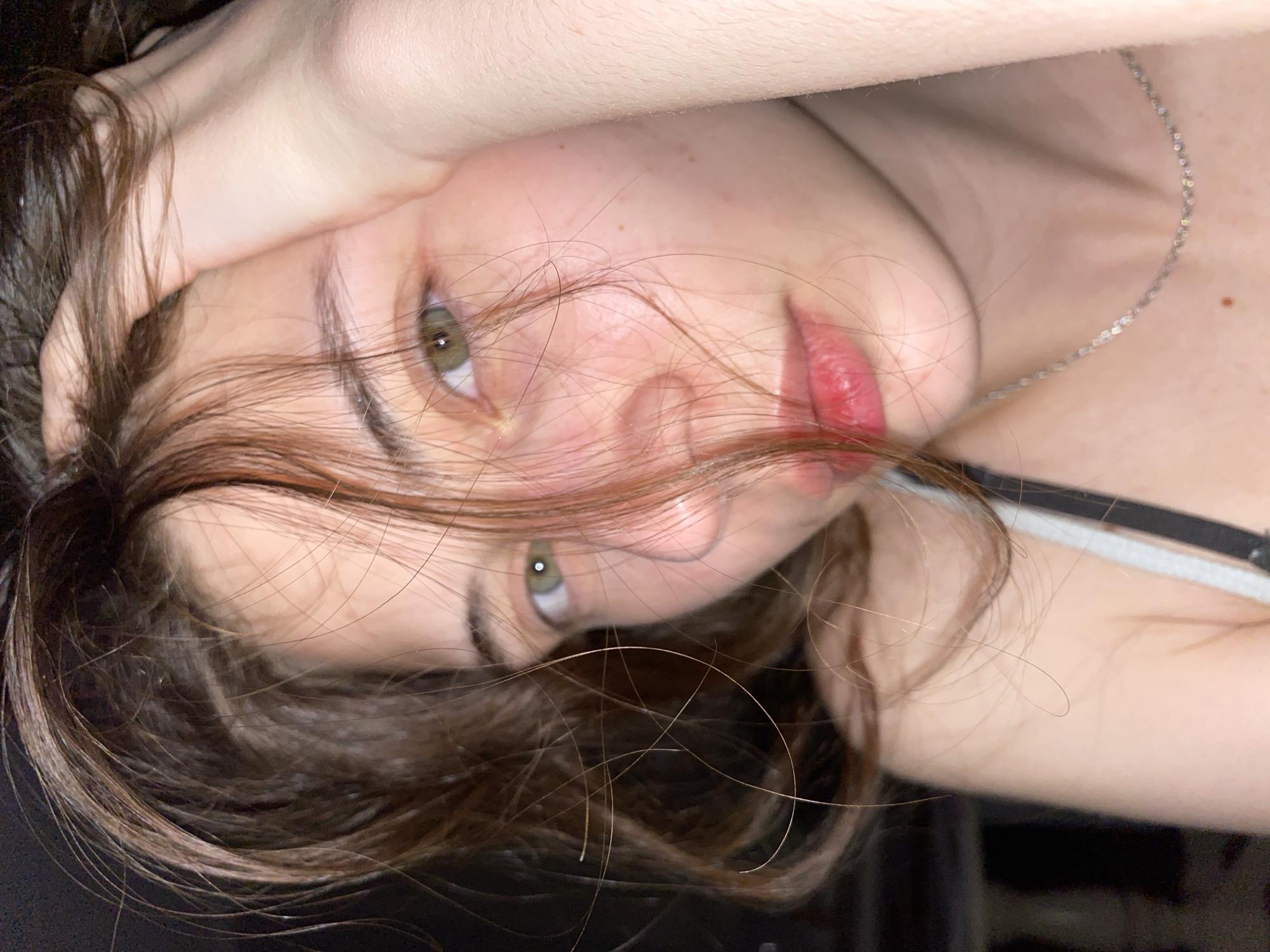 A close-up, rotated image captures a woman's face and upper body. The woman has fair skin, with visible freckles. Her eyes are green, and her lips are a shade of red. Her brown hair is partially obscuring her face. Her right hand is raised near her forehead. A thin silver necklace is visible on her neck. The background is dark. The image does not contain any information about Votkinsk, Russia.
