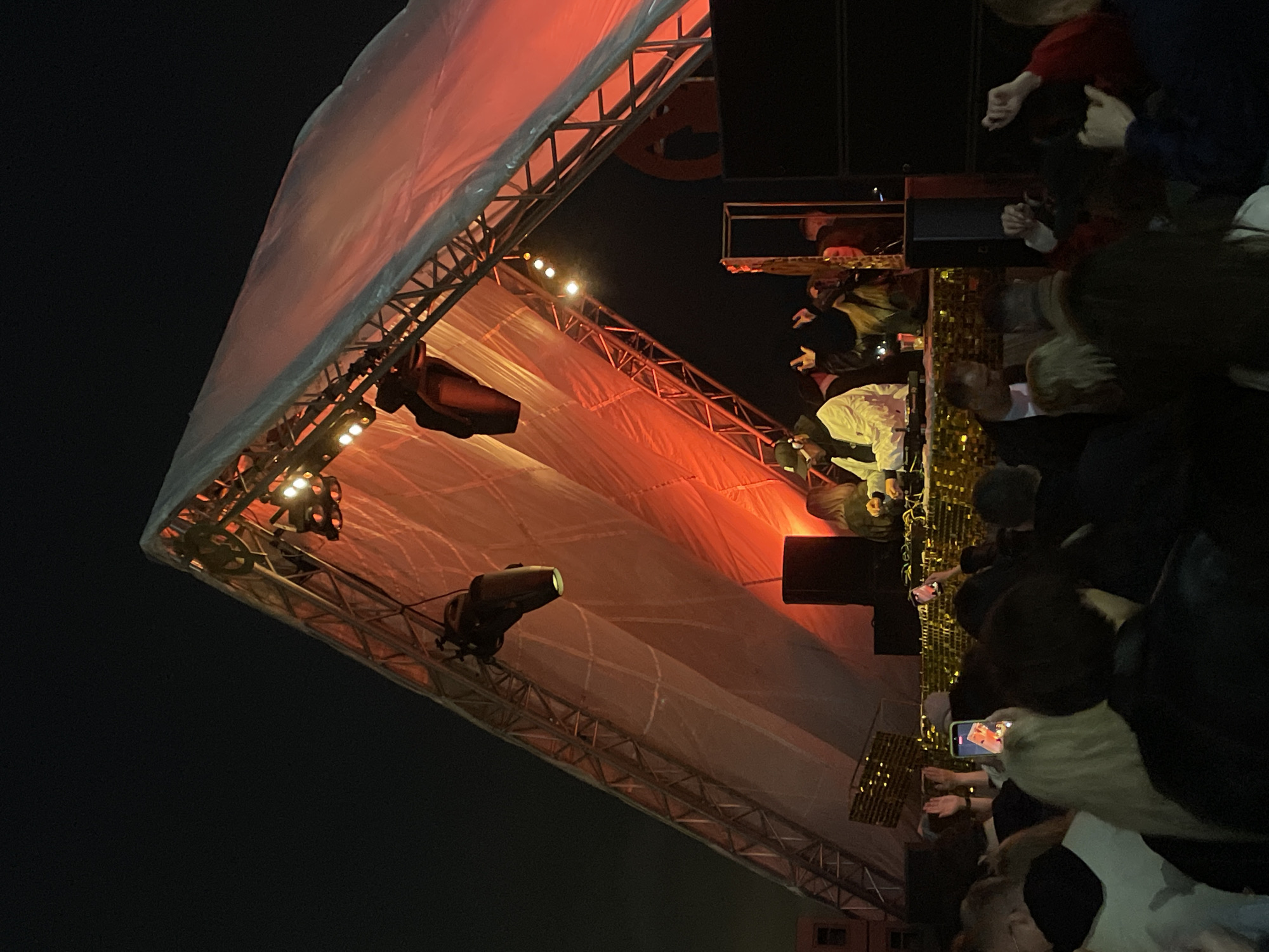 Here is a forensic description of the image:
The image depicts a concert stage with a tent-like covering, illuminated by stage lights with a reddish hue, likely at night. The stage structure appears to be made of metal trussing. Below the stage, a crowd of people is visible, some holding up phones to record or photograph the performance. The environment suggests an outdoor venue. The image provides limited information about the exact location in Saint Petersburg, Russia, but the concert setting is evident.