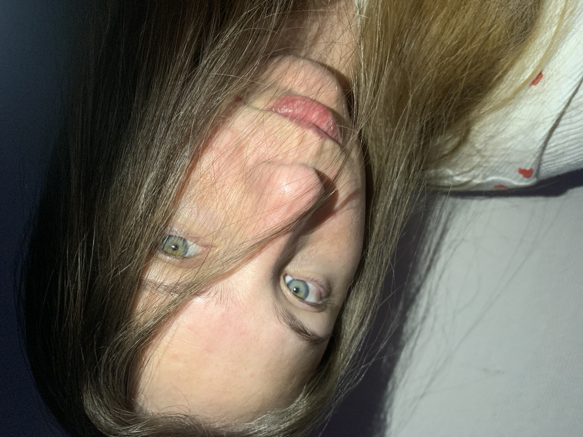 This close-up portrait features a person, likely female, with long, light brown or blonde hair, captured in a rotated orientation, appearing upside down. The subject's face is partially obscured by strands of hair, creating an intimate and somewhat mysterious atmosphere.
The person has striking green or blue eyes, which are wide open and looking directly at the viewer. Her lips are slightly parted, revealing their natural pink hue. The skin on her face appears fair, and natural skin texture is visible. On the upper right side of the image, part of a white garment with small red heart patterns can be seen beneath the hair, hinting at casual attire.
The scene is an indoor setting, characterized by strong, directional lighting. This lighting creates deep shadows on one side of the face and hair (the left side when viewed upright), contrasting with a brighter illumination on the other side. The background is simple and nondescript, dark on one side and lighter on the other, likely a wall or surface. There is no specific activity depicted other than the subject posing for the photograph. No text is visible. The time of day is not explicitly clear but the strong light suggests daylight indoors or a powerful artificial light source. The image offers no discernible location cues specific to Chaikovsky, Russia.