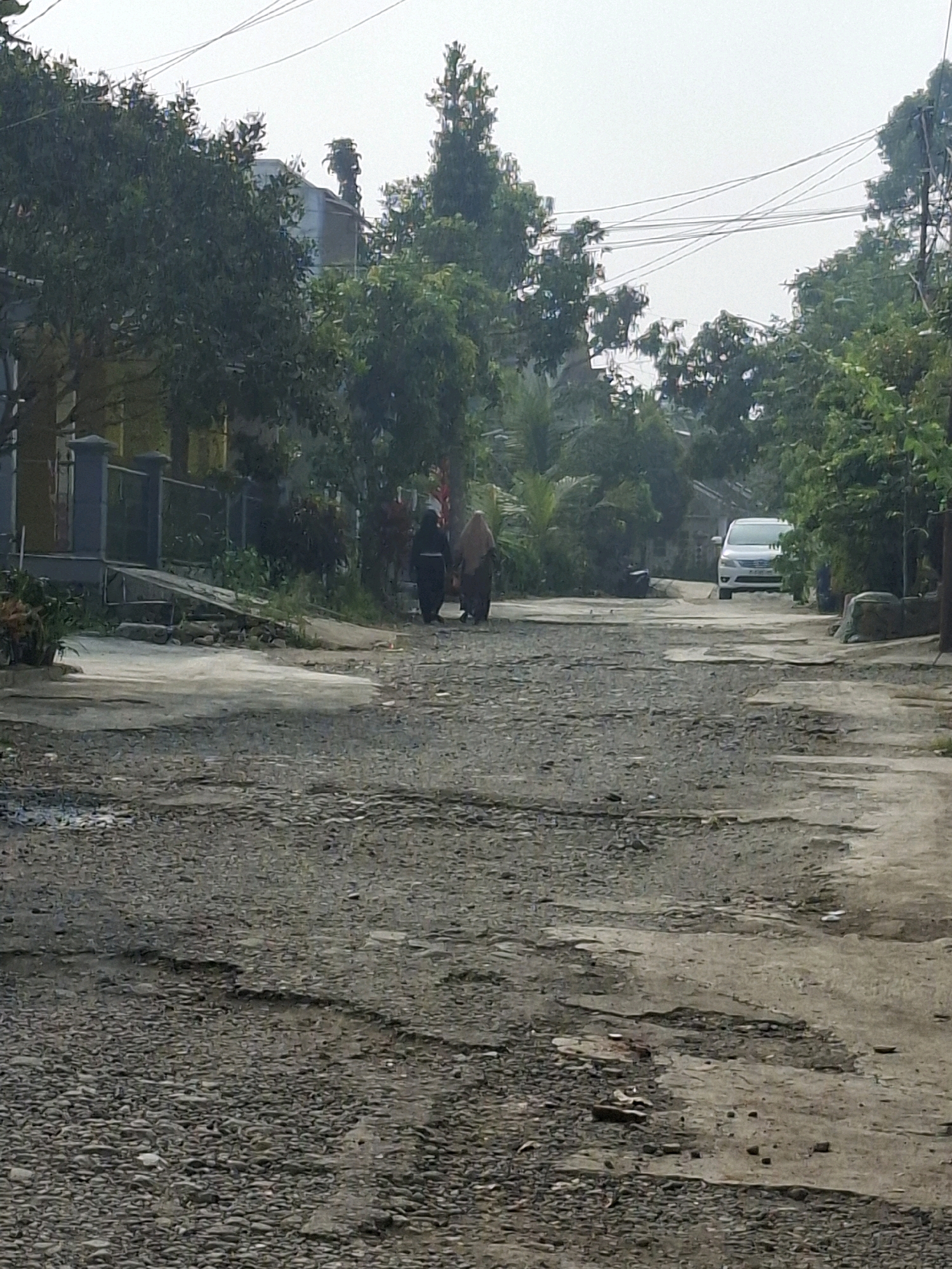The image shows a residential street in Ciheulang Tonggoh, Indonesia. The street is unpaved and appears to be in poor condition, with visible potholes and loose gravel. Several trees line the street, with dense foliage obscuring some of the houses. In the background, a white car is parked on the side of the road, and a motorcycle is visible further down. Two individuals, dressed in traditional Islamic attire, are walking away from the camera towards the car. The sky is overcast.