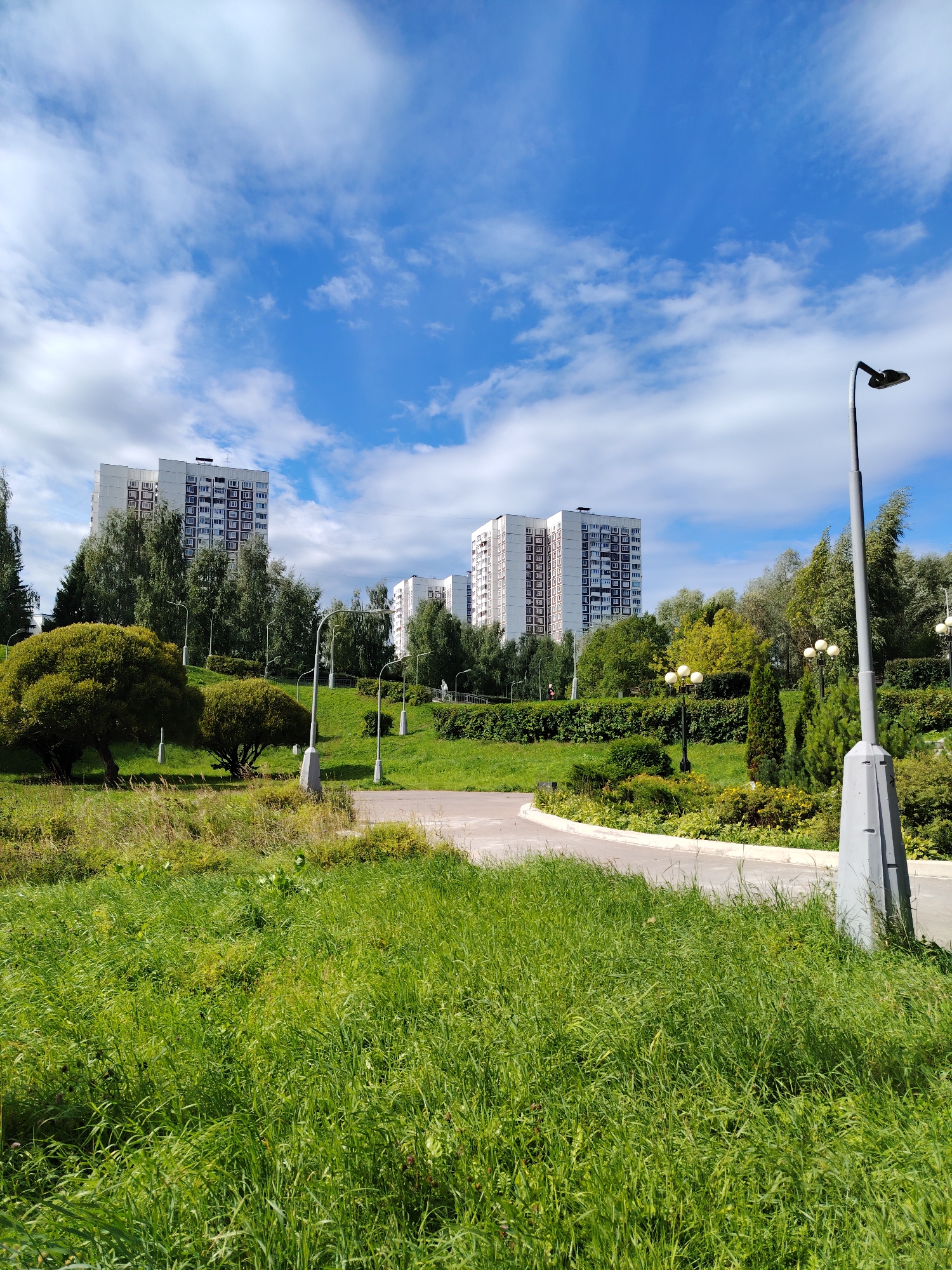 This is a sunny outdoor photograph taken in the city of Zelenograd, Russia. The image shows a park area with lush green grass in the foreground, leading to a paved pathway. Beyond the pathway, there are manicured bushes, trees, and several streetlights. In the background, two large, white apartment buildings with red-trimmed windows are visible, surrounded by a variety of trees. The sky is blue with scattered white clouds. There are no people or animals clearly visible in the image. The scene appears to be a peaceful, residential park setting.