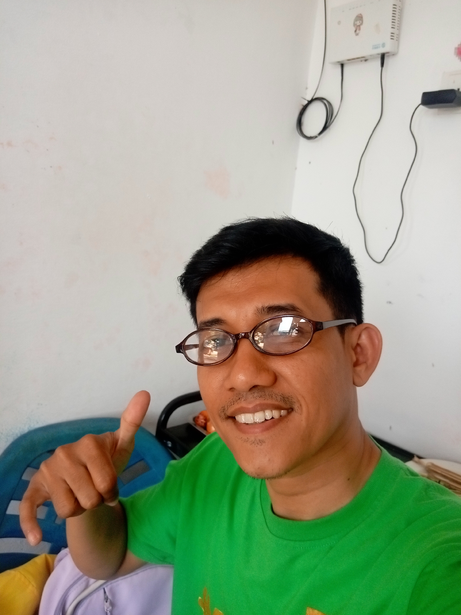 A man wearing a green t-shirt and glasses is taking a selfie in what appears to be a home or office environment. He is smiling and giving a thumbs-up gesture. Behind him, a white wall is visible with some electrical equipment mounted on it, including a white box with what looks like a cartoon character sticker and several black cables. There is also a black power adapter plugged into the wall. The man is sitting in a blue chair, and a yellow and purple bag are visible in the foreground. The overall impression is casual and informal. The location is described as Batam, Indonesia.