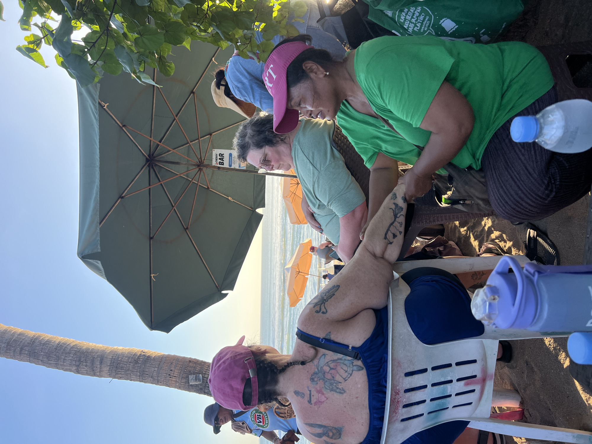 The image depicts a scene at a beachside establishment in Kuta, Indonesia. Three women are seated at tables, partially shaded by a large green umbrella. In the foreground, a woman with extensive back tattoos, wearing a blue top and a pink cap, is facing away from the camera. To her right, a woman wearing glasses and a light green shirt is seated, looking towards the ocean. Another woman, in a bright green shirt and a pink visor, is in the center right of the frame, facing the woman with tattoos and appearing to interact with her.
Behind the seated women, the ocean is visible with orange umbrellas set up on the sand. A tall palm tree is prominent on the left side of the image, with a sign that reads "CHERRY BAR" hanging from the umbrella structure. The overall setting suggests a casual dining or drinking area on the beach, with patrons enjoying the afternoon sun and ocean view. The ground is covered in sand.