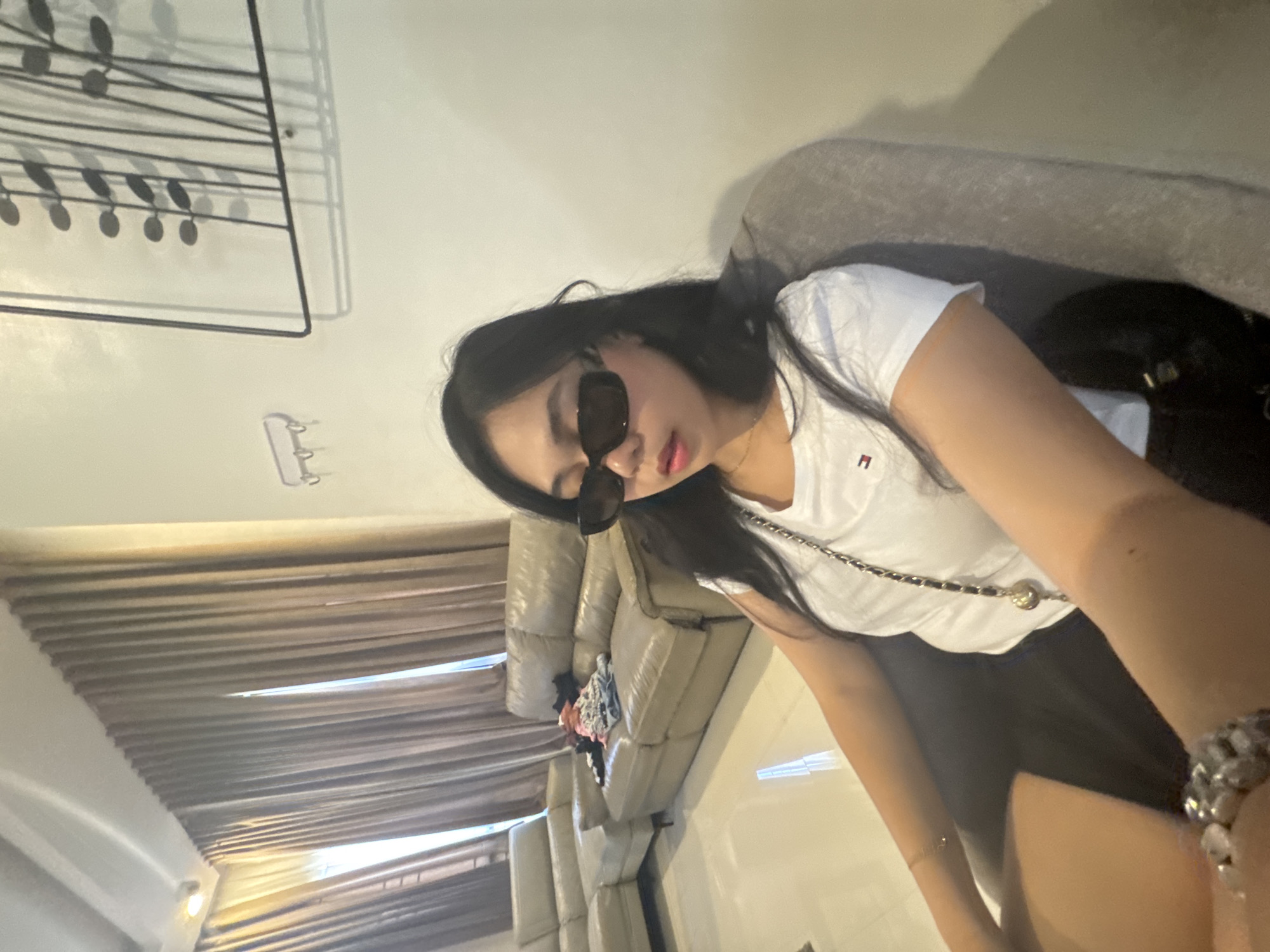 The image shows a young woman with long black hair, wearing dark rectangular sunglasses, a white t-shirt with a small red and blue logo, and a black skirt. She is seated in a grey armchair in what appears to be a living room. In the background, there is a large L-shaped beige sofa with several items of clothing piled on it. To the left, a section of a beige sectional sofa is visible. The walls are painted a light color, and there are sheer, floor-to-ceiling curtains on the left side of the frame. A piece of abstract wall art with thin black lines and circular shapes is mounted on the right wall, casting shadows. A small white key holder with three hooks is mounted on the wall to the left of the artwork. The flooring is a light-colored tile. The overall impression is of an indoor residential setting. The image does not contain specific markers to pinpoint a location within Davao City, Philippines.