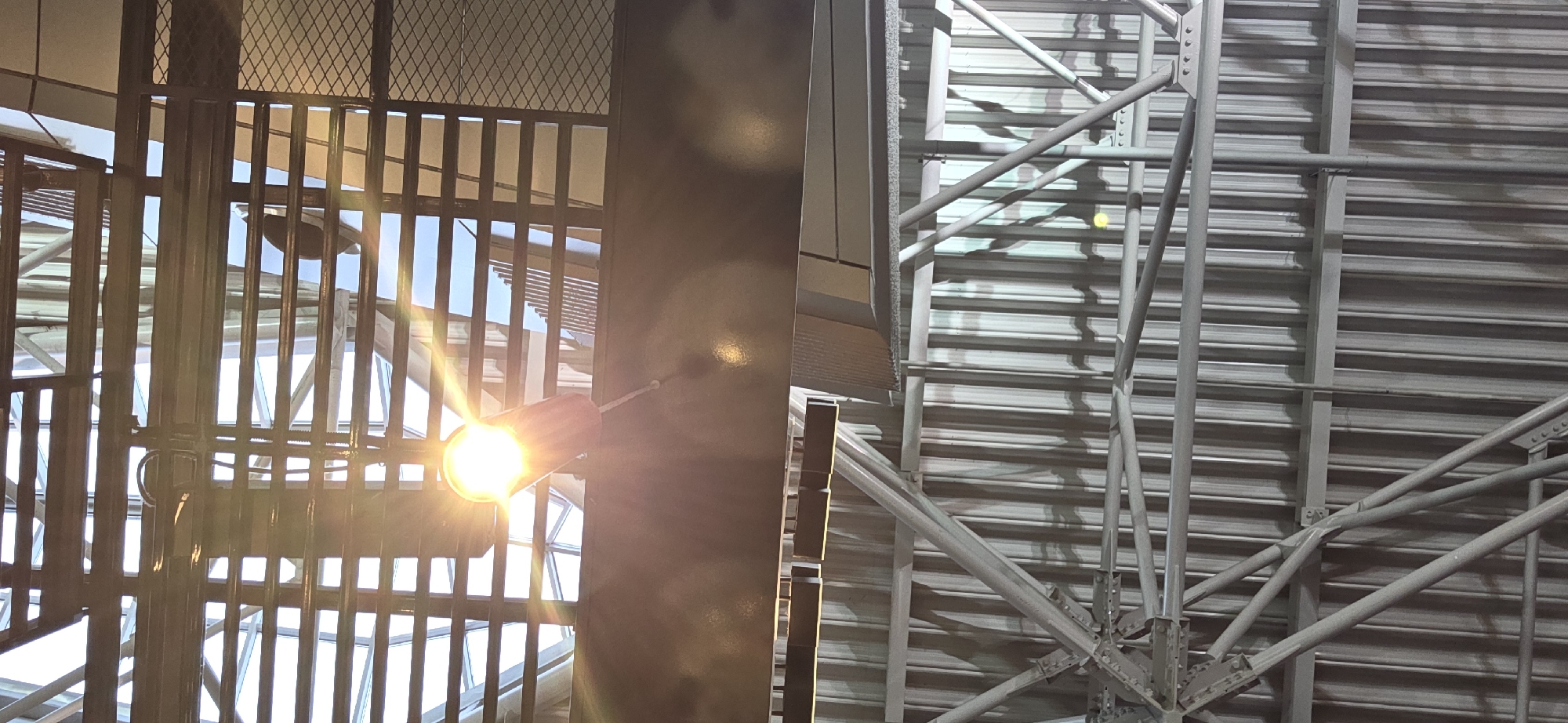 The image shows a view looking up towards the ceiling of a large, open space, likely an airport terminal, in Vnukovo, Russia. The ceiling is composed of multiple levels of industrial-style architecture. The upper level features a translucent, possibly glass, roof with a grid-like structure of dark beams. Sunlight streams through this roof, creating bright glare and lens flare from a spotlight. Below this, there is a darker, horizontal beam with reflective surfaces, upon which a black, silhouetted figure, possibly a person, is visible in the upper part of the frame. Further down, the ceiling transitions to corrugated metal panels, supported by a complex network of silver metal trusses and beams. The overall impression is one of an expansive, modern, and possibly recently constructed indoor space.