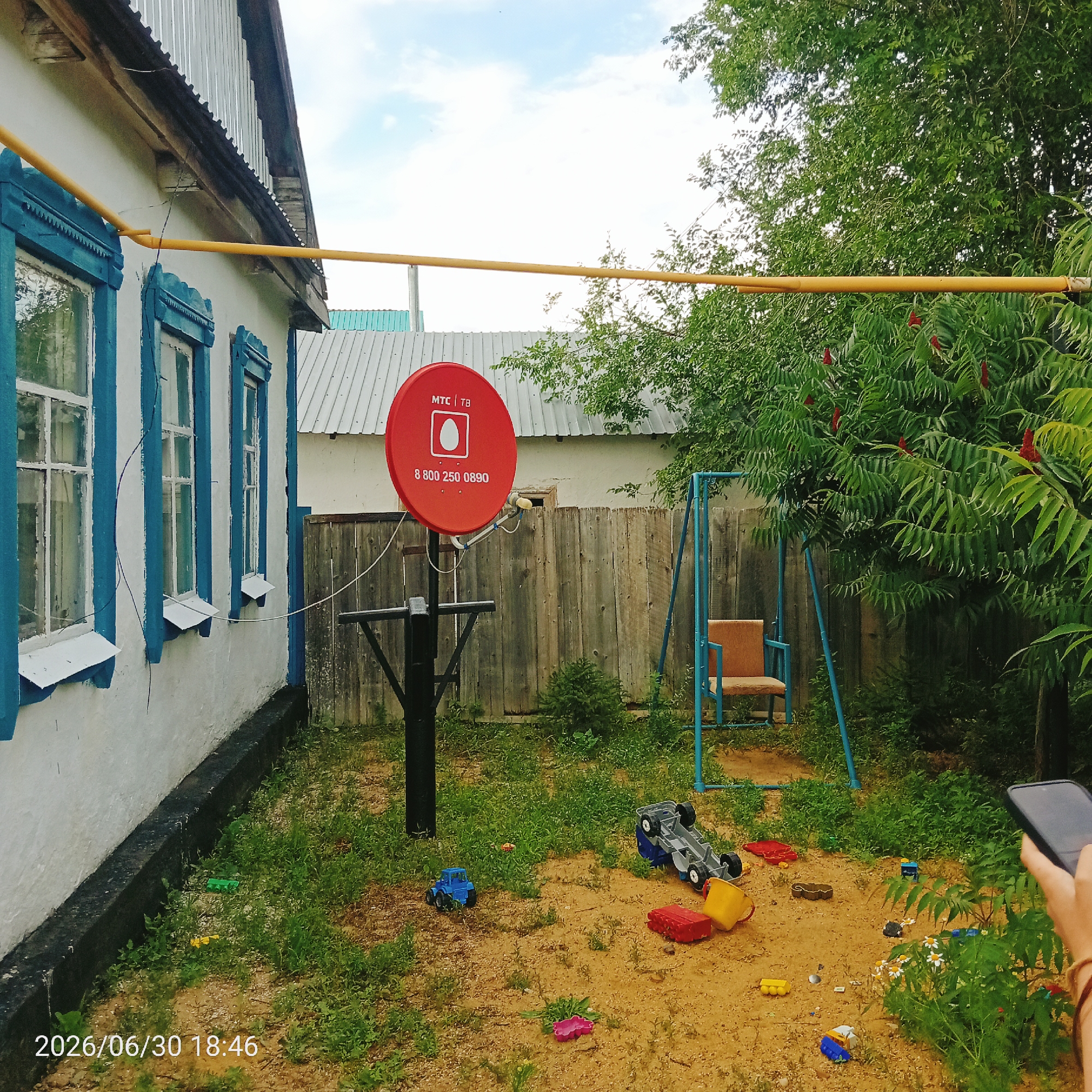 The image depicts a backyard scene, likely in a rural or suburban setting. The focal point is a red satellite dish with the MTC | TV logo and a phone number visible. The dish is mounted on a black pole in a sandy area scattered with various children's toys, including toy trucks, buckets, and building blocks. A blue swing set with an orange seat stands to the right of the dish.
In the background, a wooden fence separates the yard from more greenery. A white house with blue-trimmed windows is visible on the left side of the image, with a yellow gas pipe running along its exterior. The overall impression is one of a casual, perhaps slightly neglected, domestic outdoor space.
The time of day is indicated by the timestamp "2026/06/30 18:46" in the bottom left corner, suggesting late afternoon or early evening. The sky is partially cloudy, indicating fair weather. The presence of children's toys suggests the area is used for play, though no people are directly visible in the frame. A hand holding a smartphone is visible on the far right, hinting at someone taking the photograph.