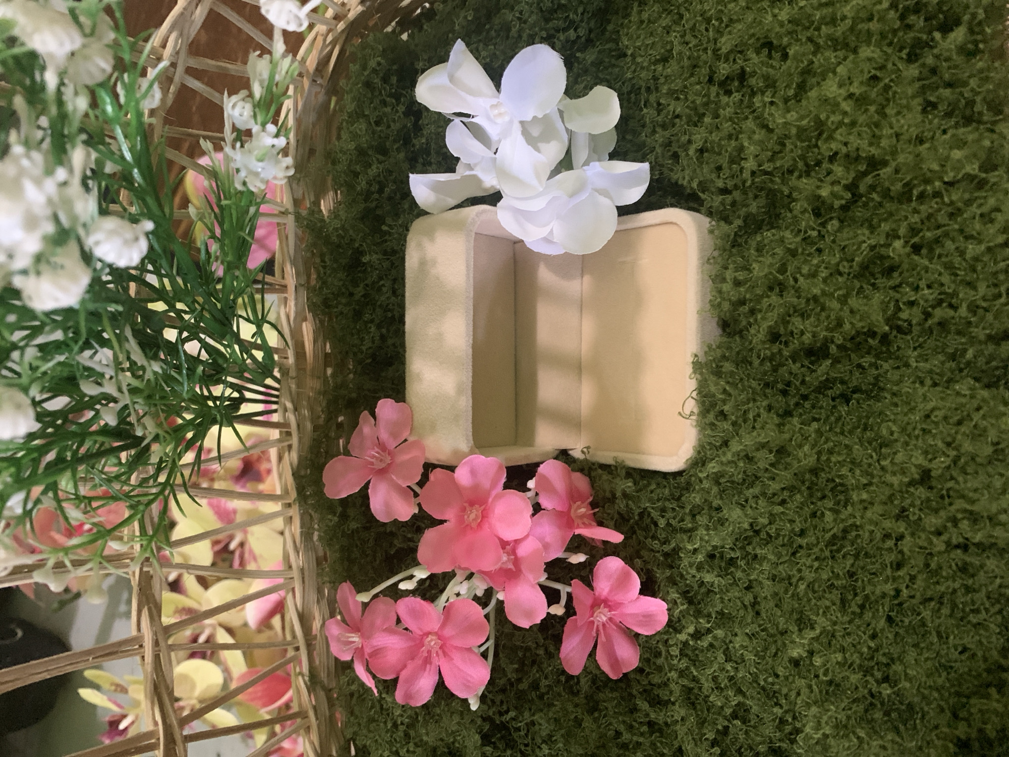 The image displays a close-up, eye-level shot of a rustic arrangement within a woven basket. The foreground is dominated by a thick layer of dark green, textured material resembling moss or artificial grass, upon which a single, open, beige velvet ring box is centrally placed. To the left of the box, a cluster of five vibrant pink artificial flowers is arranged. To the right of the box, a small bunch of white artificial flowers with pearl-like centers is positioned. In the background, partially obscured by the foreground elements and the basket's weave, are artificial flowers in shades of yellow, pink, and white, along with green foliage. The lighting is soft and even, casting no distinct shadows. There are no people visible in the image, nor any discernible urban elements to suggest a specific location within Kuala Pembuang, Indonesia. The scene is static and appears to be a still life arrangement.