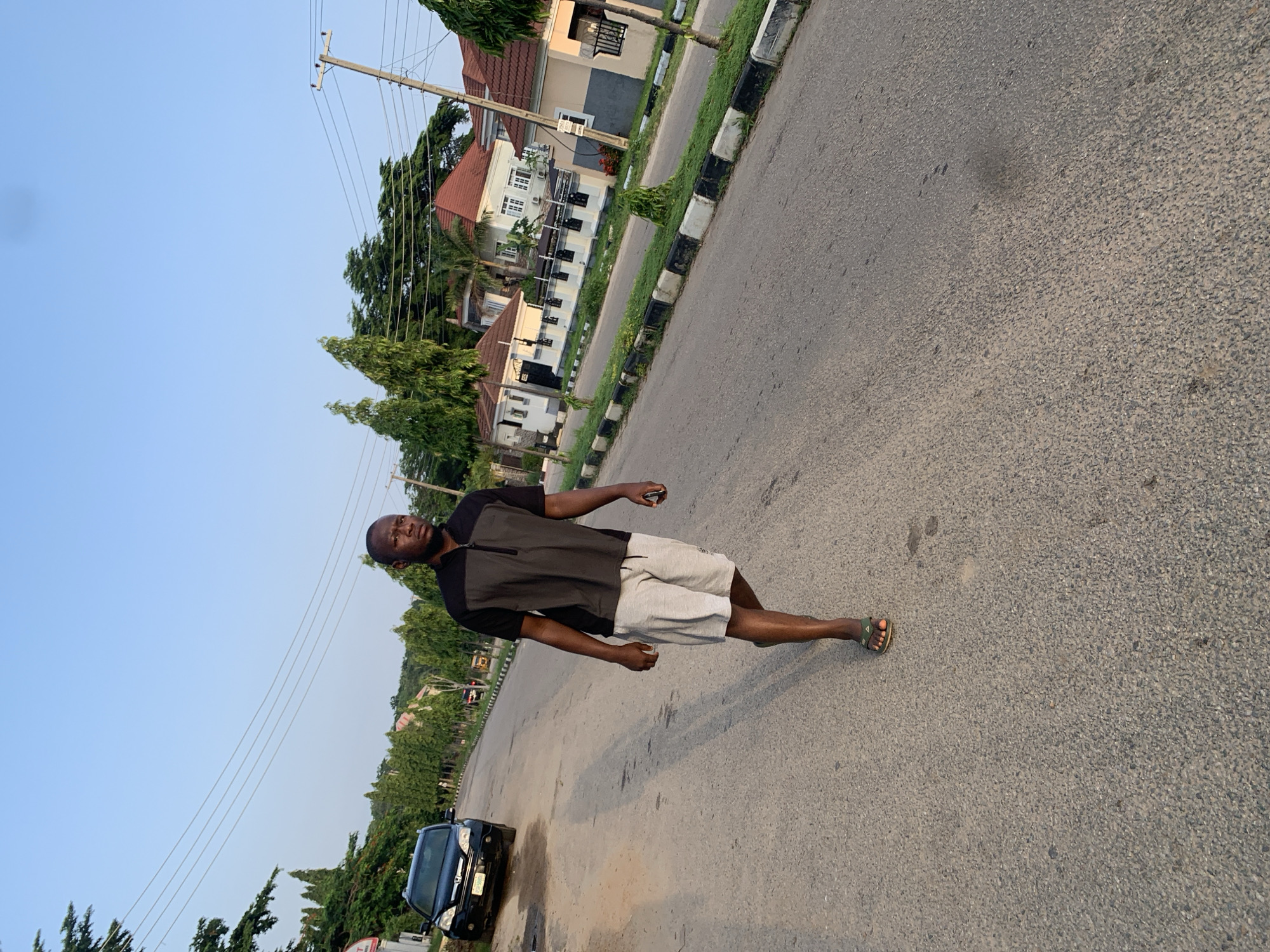 A full shot photograph captures a Black man walking on a paved road. He is wearing a black short-sleeved shirt with a gray zipper and grey shorts. He has on a pair of green sandals. The man is posing as he walks, looking towards the camera with a neutral expression. The road is asphalt with a painted white and black curb. In the background, there are several houses with red tile roofs, lush green trees, and utility poles with wires. A black car is parked on the side of the road to the left. The scene is outdoors, with a clear blue sky visible above. The lighting suggests it is either early morning or late afternoon, as the sun casts a shadow of the man on the road. The overall impression is a casual, everyday scene in a residential area. The location context is Abuja, Nigeria. There is no visible text in the image.