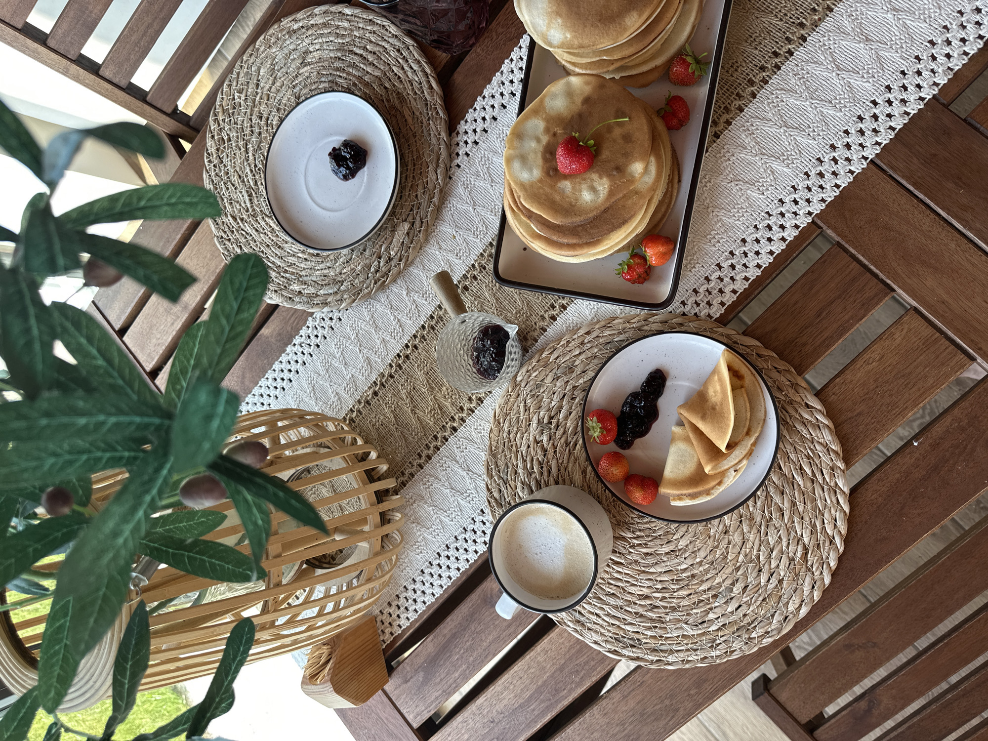 The image depicts a close-up, overhead view of a wooden table set for breakfast. On the table, a woven placemat supports a white mug of coffee with latte art. To the right of the mug, another woven placemat holds a white plate with folded pancakes, a dollop of dark jam, and three fresh strawberries. Further to the right, a rectangular platter overflows with a stack of golden-brown pancakes, garnished with a single strawberry on top and surrounded by several more strawberries. A small glass dish containing jam is also visible. In the background, a decorative basket with artificial olive branches and olives is positioned on the table. The scene is framed by the slats of a wooden chair. The overall ambiance suggests a relaxed, outdoor breakfast setting, possibly on a balcony or patio, given the glimpse of greenery outside and the natural light. The location is identified as Самарский, Russia.