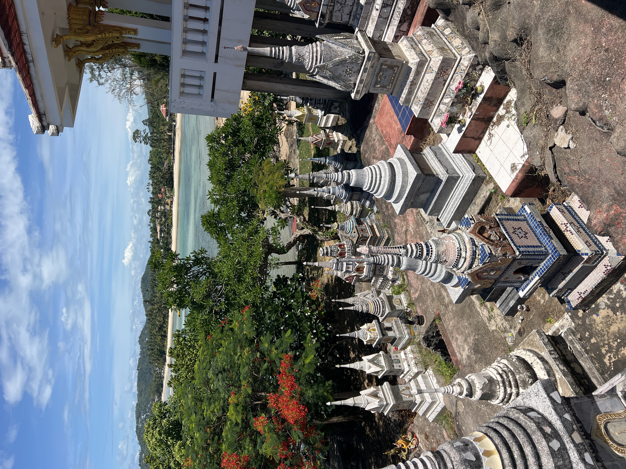The image displays an elevated view of a coastal scene in Baan Hua Thanon, Thailand, characterized by a unique array of structures in the foreground.
In the immediate foreground, numerous small, spire-topped monuments are densely arranged on tiered concrete and dirt platforms. These structures, resembling miniature stupas or ossuaries, vary significantly in design and material. Many are constructed from light gray concrete, featuring tiered bases and pointed finials. Others exhibit intricate decorative patterns, incorporating mosaic tiles in shades of blue, pink, and white, with some featuring gold accents. Several monuments show signs of weathering, with discoloration and surface degradation. On one of the tiered platforms, two small vases containing pink flowers are present, indicative of offerings.
To the right, a portion of a white building with a red-tiled roof is visible, supported by white pillars, and featuring an ornate gold support structure and a white balustrade extending from a balcony. The ground surrounding the monuments is a mix of weathered concrete, exposed earth, sparse weeds, and natural rock formations.
The midground is dominated by lush tropical vegetation. A prominent tree on the left features dense green foliage interspersed with clusters of bright red-orange flowers. Other trees display rich green leaves. Beyond this vegetation, a calm, clear turquoise-blue bay stretches across the image, transitioning to deeper blue further from shore. A narrow, light sandy beach runs along the bay's edge. A small yellow boat is visible on the water in the mid-distance.
In the background, a series of verdant green hills rise, covered with dense foliage and dotted with scattered buildings, presumably residential or resort properties. The sky above is a bright blue, accented by scattered white cumulus clouds. No human figures are visible within the frame.
