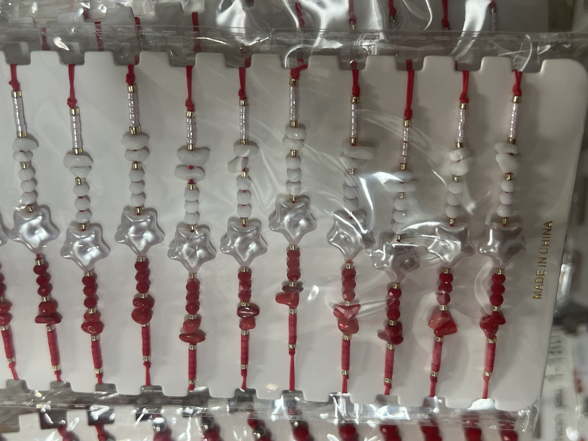 A close-up shot shows several identical good luck charms packaged in clear plastic. Each charm features a red string adorned with small red and white beads and a star-shaped clear or white bead near the center. The packaging is white, and "MADE IN CHINA" is printed on the right side. The charms are arranged in rows. The scene is most likely inside a shop, possibly in the city of Burgas, Bulgaria.
