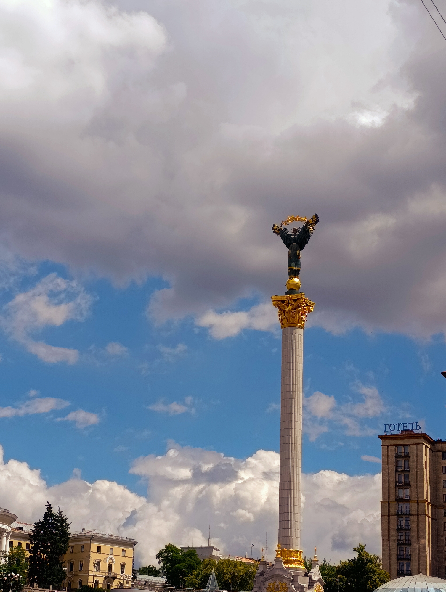 This is a medium shot, taken outdoors on a cloudy day, of the Independence Monument in Kyiv, Ukraine. The monument, a tall column topped with a statue of a woman holding a laurel wreath, dominates the center of the frame. The sky above is a mixture of bright blue with fluffy white clouds and darker, more ominous gray clouds. To the right of the monument, a tall, multi-story building with many windows is visible. A sign on top of this building reads "ГОТЕЛЬ" in blue Cyrillic letters, indicating it is a hotel. In the foreground and to the left, glimpses of other buildings and trees can be seen. The overall scene suggests a bustling urban environment, likely a public square or plaza.