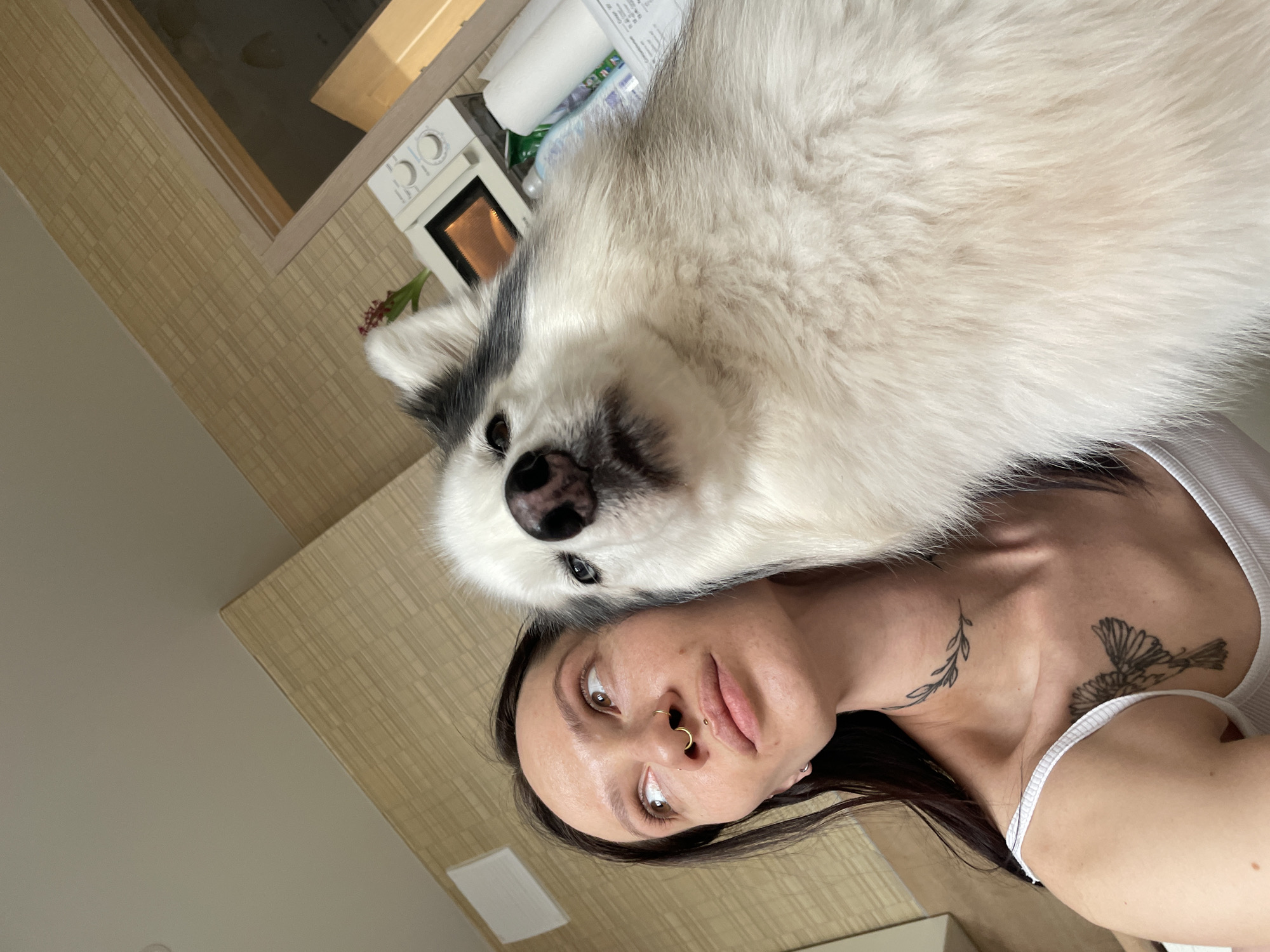 An indoor scene depicts a female subject and a dog. The female, with dark hair, a gold septum piercing, a labret piercing, a vine-like neck tattoo, and a bird tattoo on her right chest, is wearing a white tank top. She is looking upwards and to the left of the frame. Adjacent to her head is a large canine, identified as a Husky or Malamute type, with white and grey fur, a black nose, and an apparent blue eye. The dog's head is directed towards the camera. The background consists of a textured, light brown patterned wallpaper and a plain light-colored ceiling. To the upper right, a white microwave oven is visible on a surface, with a red floral object and other miscellaneous items. A wooden door frame is partially visible in the far right background. The specific location within Izhevsk, Russia, is not identifiable from the provided visual information, but the environment suggests a residential interior.