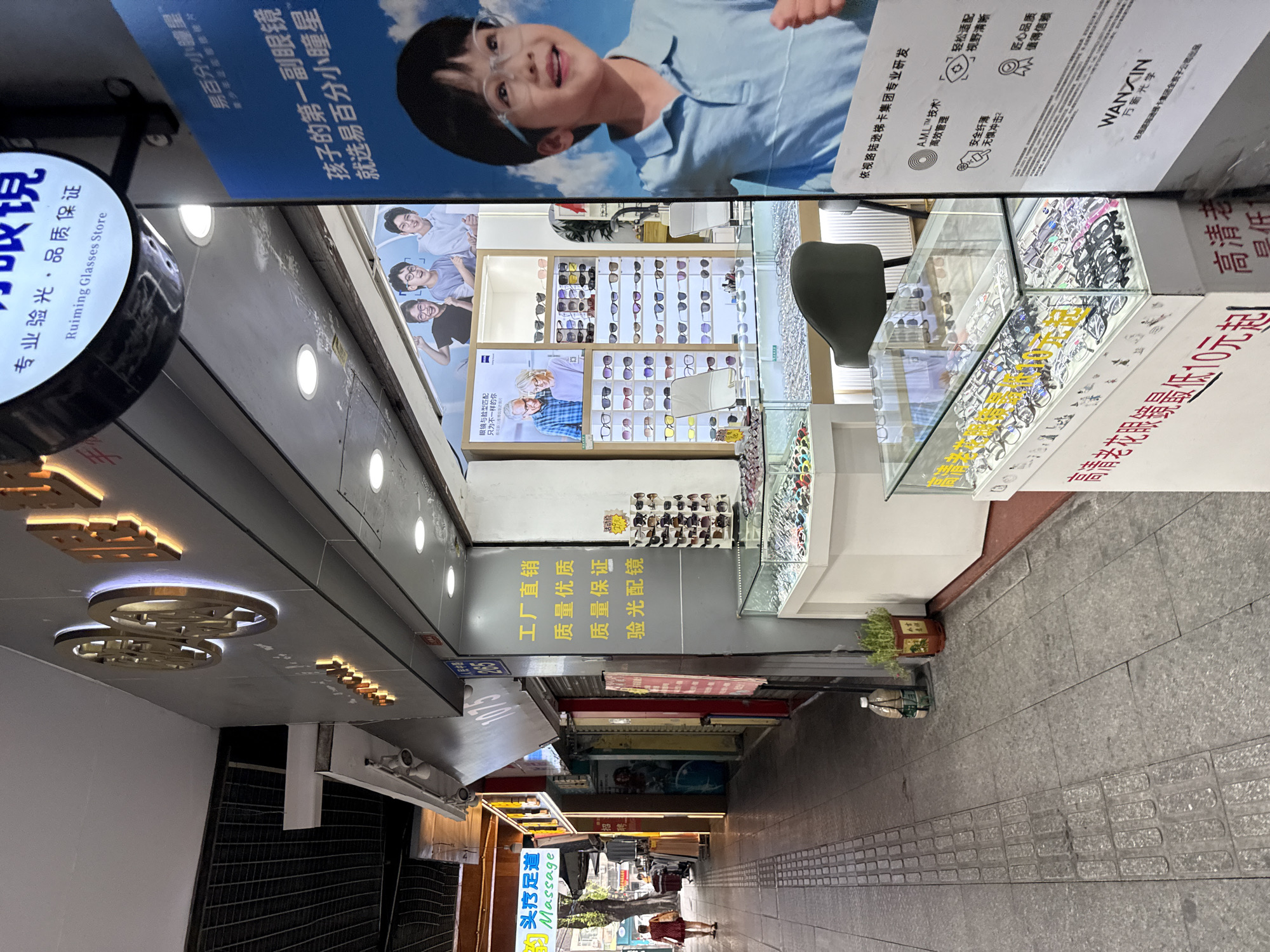 The image shows a street-level view of an optical shop in Yuexiu District, China. The shop's facade is illuminated by bright signage, with large golden characters and a circular logo that reads "Ruiming Glasses Store" in English. Inside, the shop displays a variety of eyeglasses and sunglasses on shelves and in glass cases. A large poster features a young boy wearing glasses, and other advertisements are visible. The street outside is paved with grey tiles, and a woman in a red dress is walking away from the camera. The overall scene depicts a typical commercial street in an urban Chinese setting, with a focus on retail and consumer goods.
