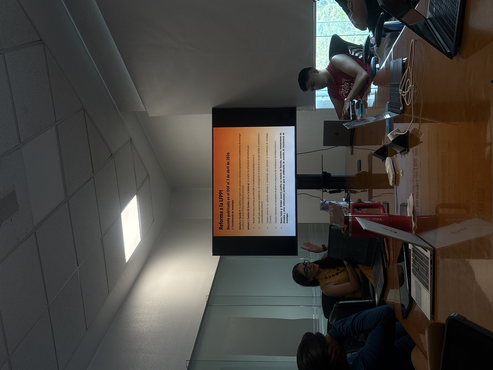 Here's a description of the image:
**Setting:** The image appears to be taken inside an office or conference room. There is a long wooden conference table with laptops and other items on it. A large screen is mounted on the wall, displaying a presentation slide in Spanish about reforms to the "LFPPI" (Ley Federal de Protección de la Propiedad Industrial) published in the Diario Oficial de la Federación (DOF) on April 3, 2026, focusing on technology transfer.
**People:** Several people are present around the table. They are engaged in what seems like a meeting or presentation. One woman is gesturing towards the screen, seemingly explaining the content. Another person is visible in the foreground, and there's a man wearing a red shirt in the background.
**Objects:** The main objects in the scene include:
* Laptops
* Conference table
* Mounted presentation screen
* Chairs
**Actions/Interactions:** People are seated around the table, looking at the screen, and engaged in discussion.
**Location Information:** Puebla, Mexico can be inferred from the context and presentation content.