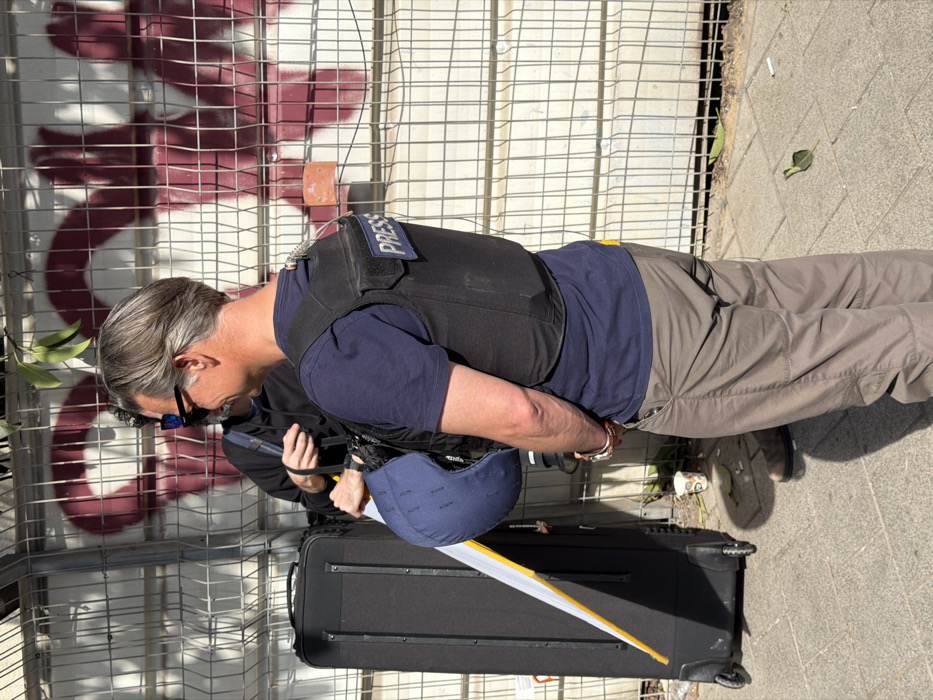 The image displays two males standing on a paved sidewalk in Tel-Aviv, Israel, under direct sunlight. The foreground subject, a male with grey hair, is seen from the mid-back and right profile. He wears a dark blue short-sleeved t-shirt, light brown cargo pants, and a black tactical vest bearing a "PRESS" patch on the upper back. An electronic device with a clear cable is attached to the vest. He holds a dark blue cap with an attached white and yellow protective visor in his left hand, while multiple silver bracelets adorn his right wrist.
Behind him and to the left, partially obscured by a black wheeled suitcase, stands a second male. This individual wears a black hooded sweatshirt and dark sunglasses and holds a black item with a strap.
The background features a grey metal grid fence, behind which a solid surface is covered with large, abstract red/maroon graffiti. The paved sidewalk shows a white disposable cup, a green leaf, and a cigarette butt. Green leaves from an overhead branch are visible at the top of the frame, casting distinct shadows on the ground.