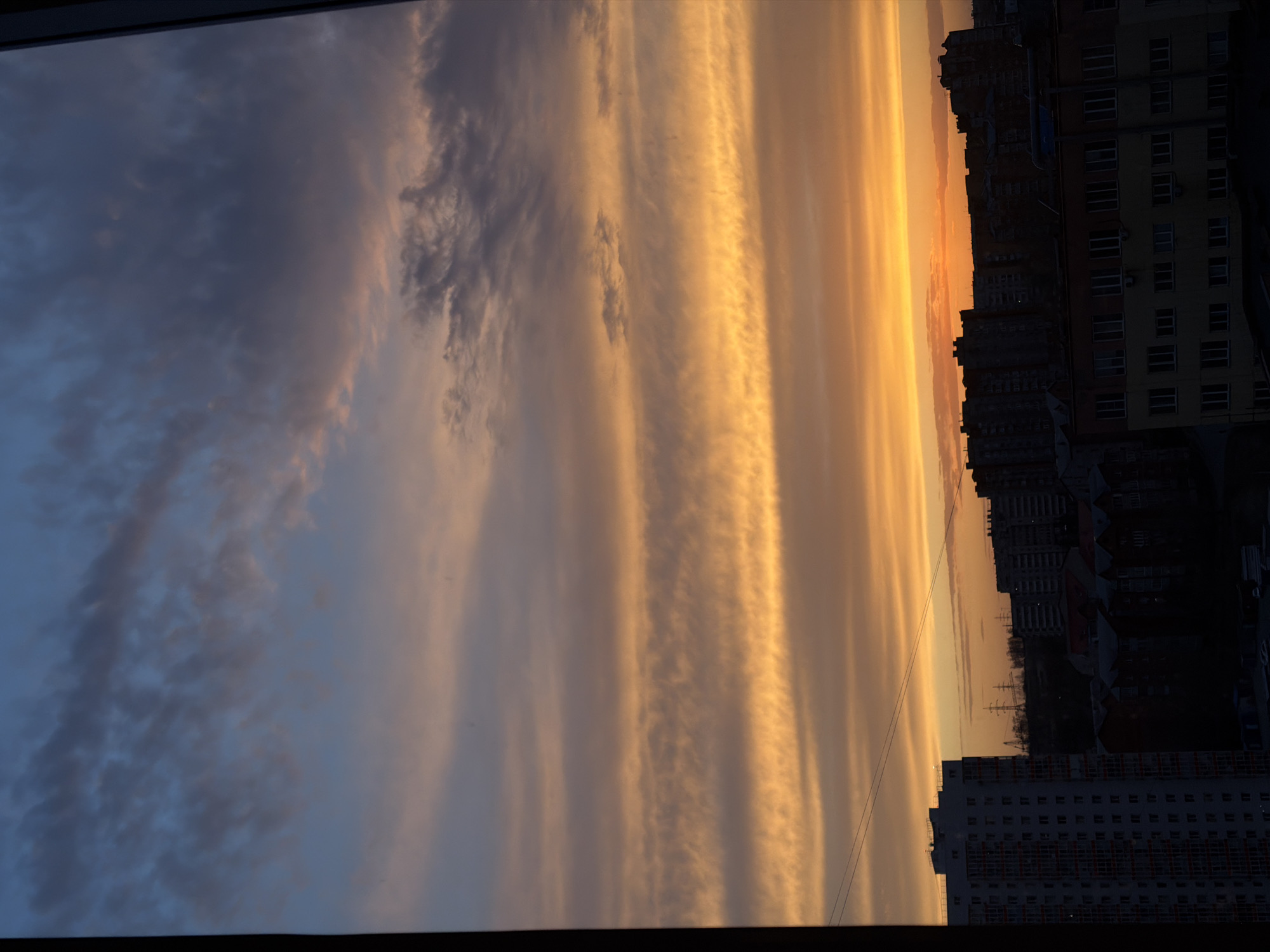 The image shows a view from a high vantage point, possibly a window in a building. The sky dominates the scene, exhibiting a sunset with layered clouds. The clouds transition from a pale blue-gray on the left to a bright orange and yellow near the center and right, indicating the setting sun. Buildings are visible on the right side of the image, silhouetted against the sky. One building in the foreground is light-colored with many windows. There are also power lines in the sky to the right. The location of this photo is the city of Krasnoyarsk, Russia.