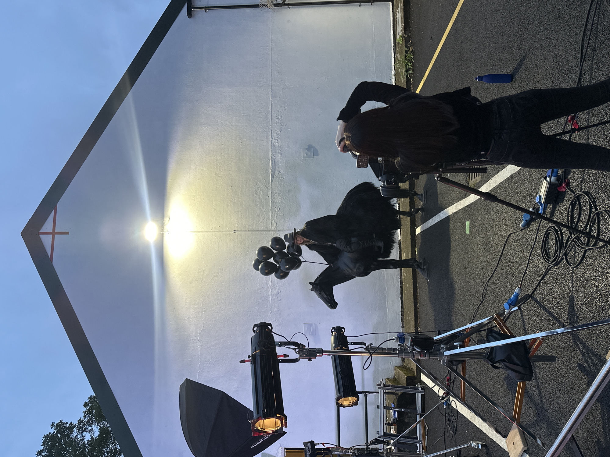A staged outdoor photoshoot is taking place in front of a white wall. A person dressed in dark clothing and a hat is seated on a black, faux-fur horse. The person is holding a cluster of black balloons. Several professional lighting rigs are set up around the scene, including a large softbox and two spotlight-style lights. A photographer, viewed from behind, is actively taking pictures of the subject with a camera mounted on a tripod. The ground appears to be asphalt, suggesting an urban or suburban setting. The sky is overcast and dark, indicating it is either dusk or a heavily clouded day. The building in the background has a white facade and a dark-pitched roof with a red cross emblem at its peak, hinting at a church or similar institution. This scene is taking place in London, United Kingdom.