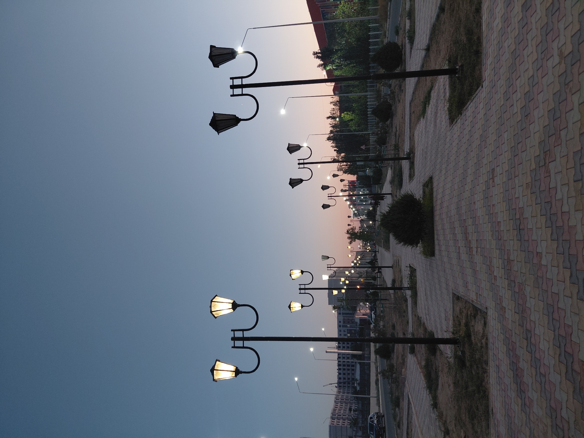 This image depicts a street scene in Aktau, Kazakhstan at dusk. A paved walkway, made of colorful interlocking bricks, runs parallel to a road. Several ornate lampposts with multiple glowing bulbs line the walkway, illuminating the path. On the left side of the road, there are buildings under construction and parked cars. The sky transitions from a deep blue at the top to shades of pink and orange near the horizon, indicating the setting sun. The overall environment is one of urban development transitioning into evening.